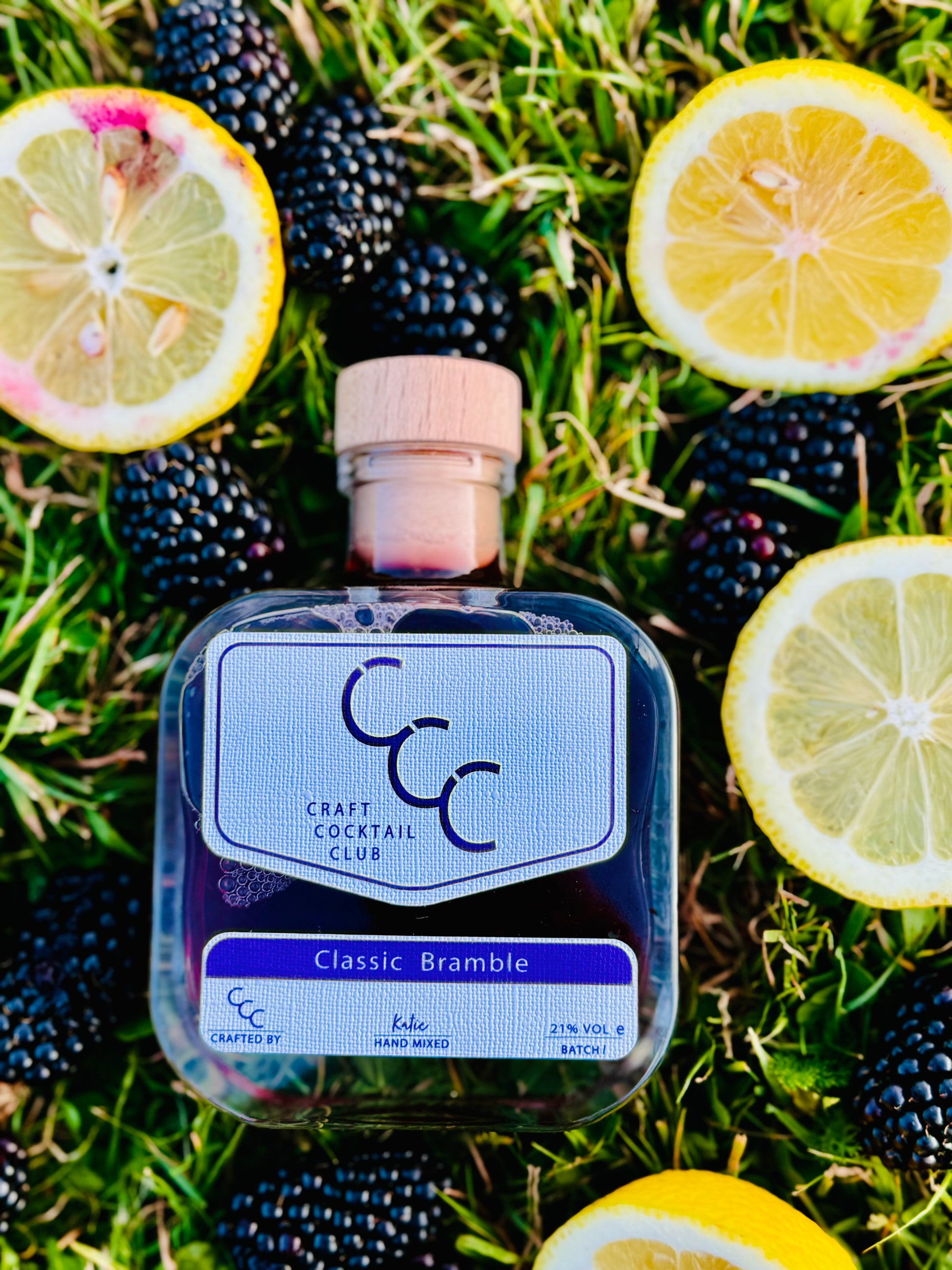 Picture of a bottle lying flat in the grass surrounded by juicy blackberries and cut lemons, the label on the bottle reads Craft cocktail club, classic Bramble 