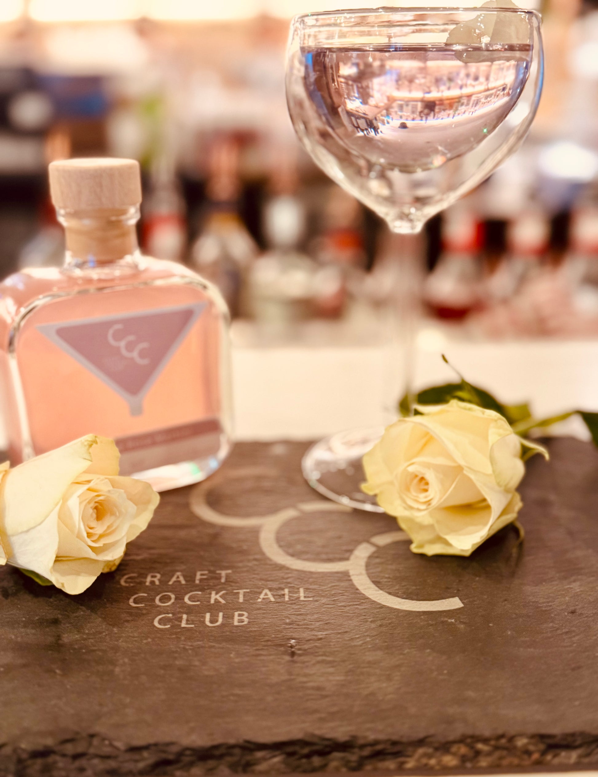 Picture of a pink cocktail on a dark slate dial with white roses, next to the glass is a square glass bottle with pre mixed cocktail inside label reads craft cocktail club 