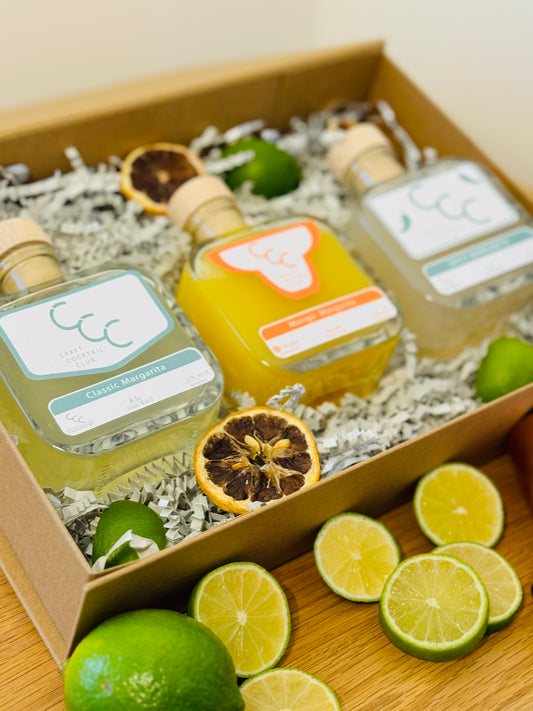 picture of a gift box with three margaritas  inside, the bottles a green and orange and. the box is surrounded by green fruit 