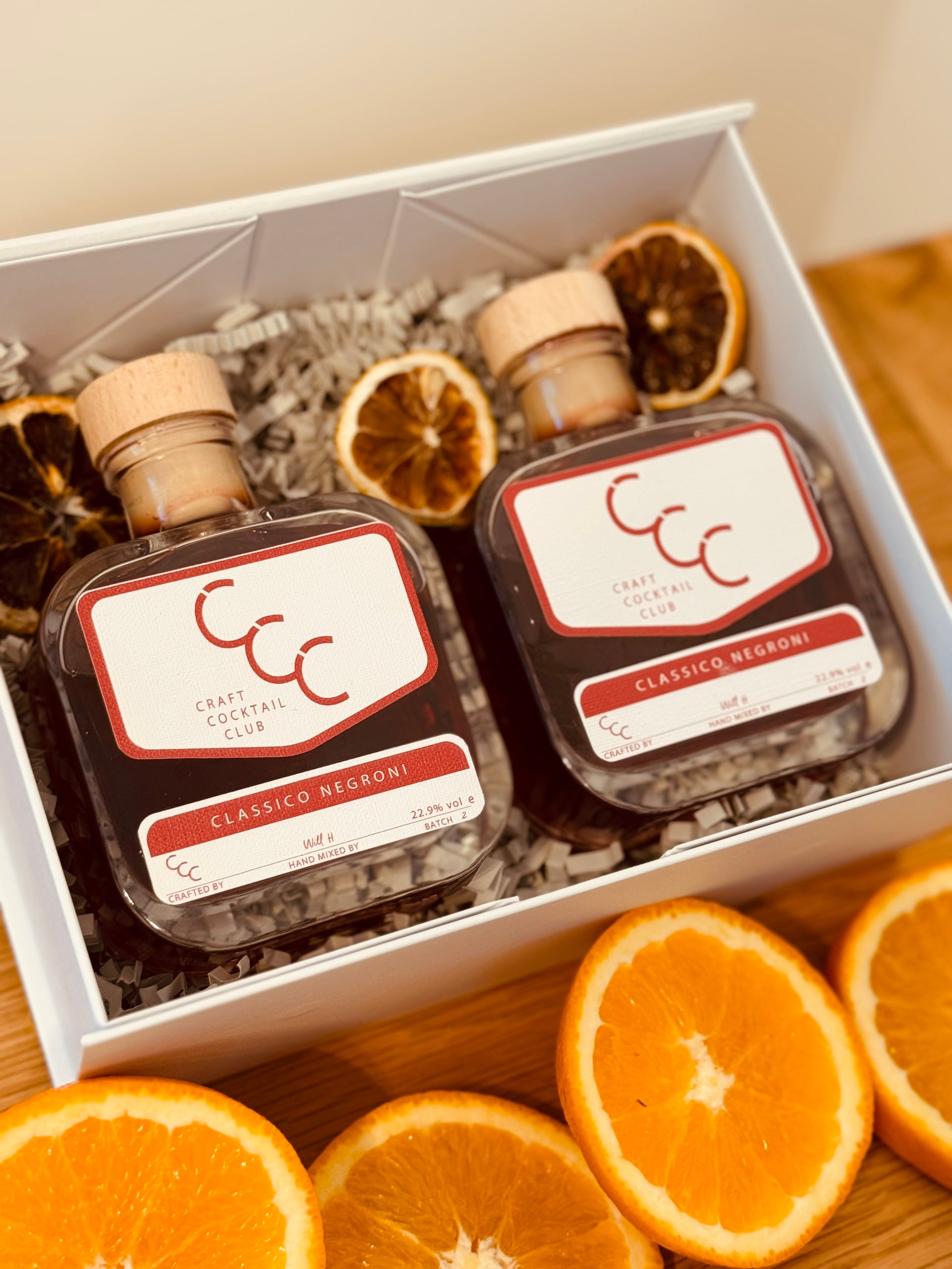 Picture of a gift box surrounded by oranges, in the gift box are two glass bottles full of dark red liquid, the labels read craft cocktail club Classic Negroni 