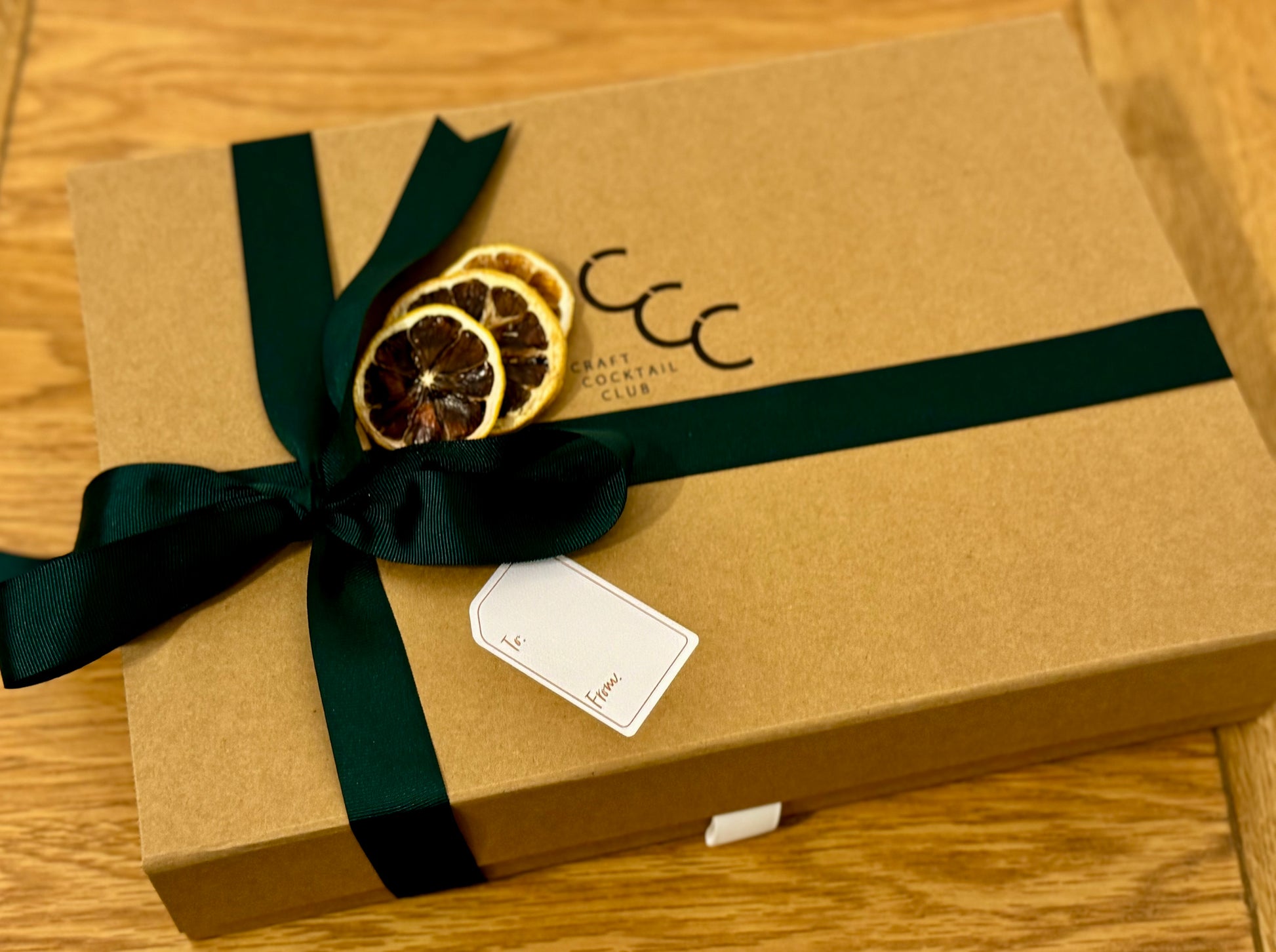 picture of a brown gift box with a green ribbon wrapped around it. dried fruit rest on to and he logo says craft cocktail club 