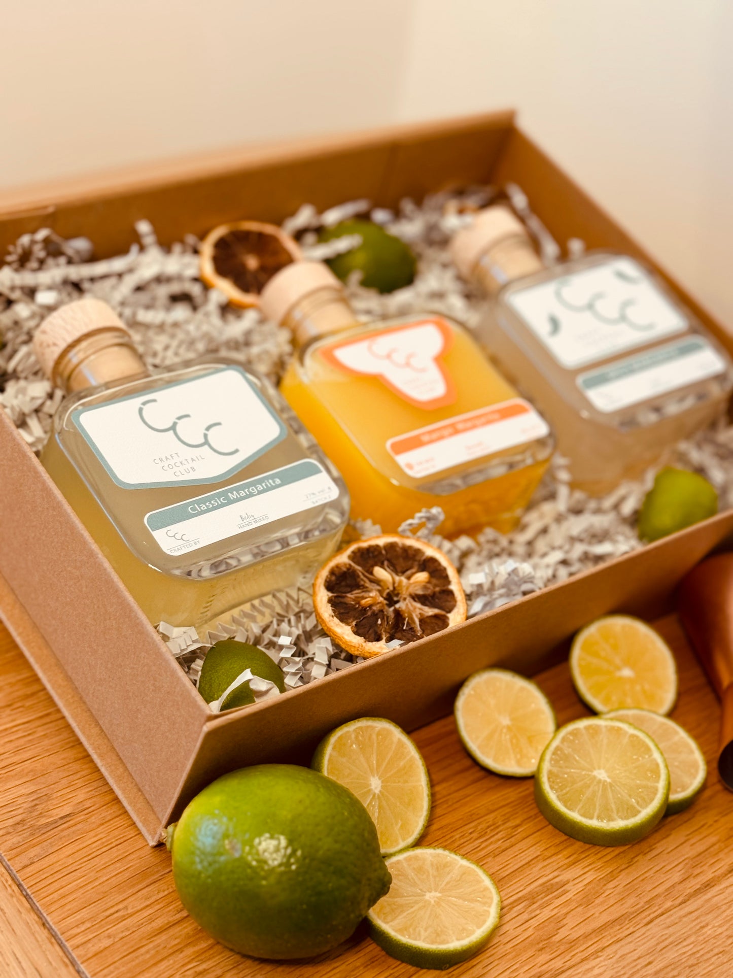 picture of a gift box with three margaritas  inside, the bottles a green and orange and. the box is surrounded by green fruit 