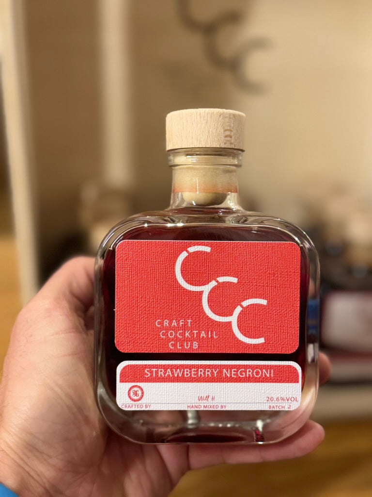 Picture of a square bottle n a mans hand,  it has a red label that reads craft cocktail club, strawberry negroni 