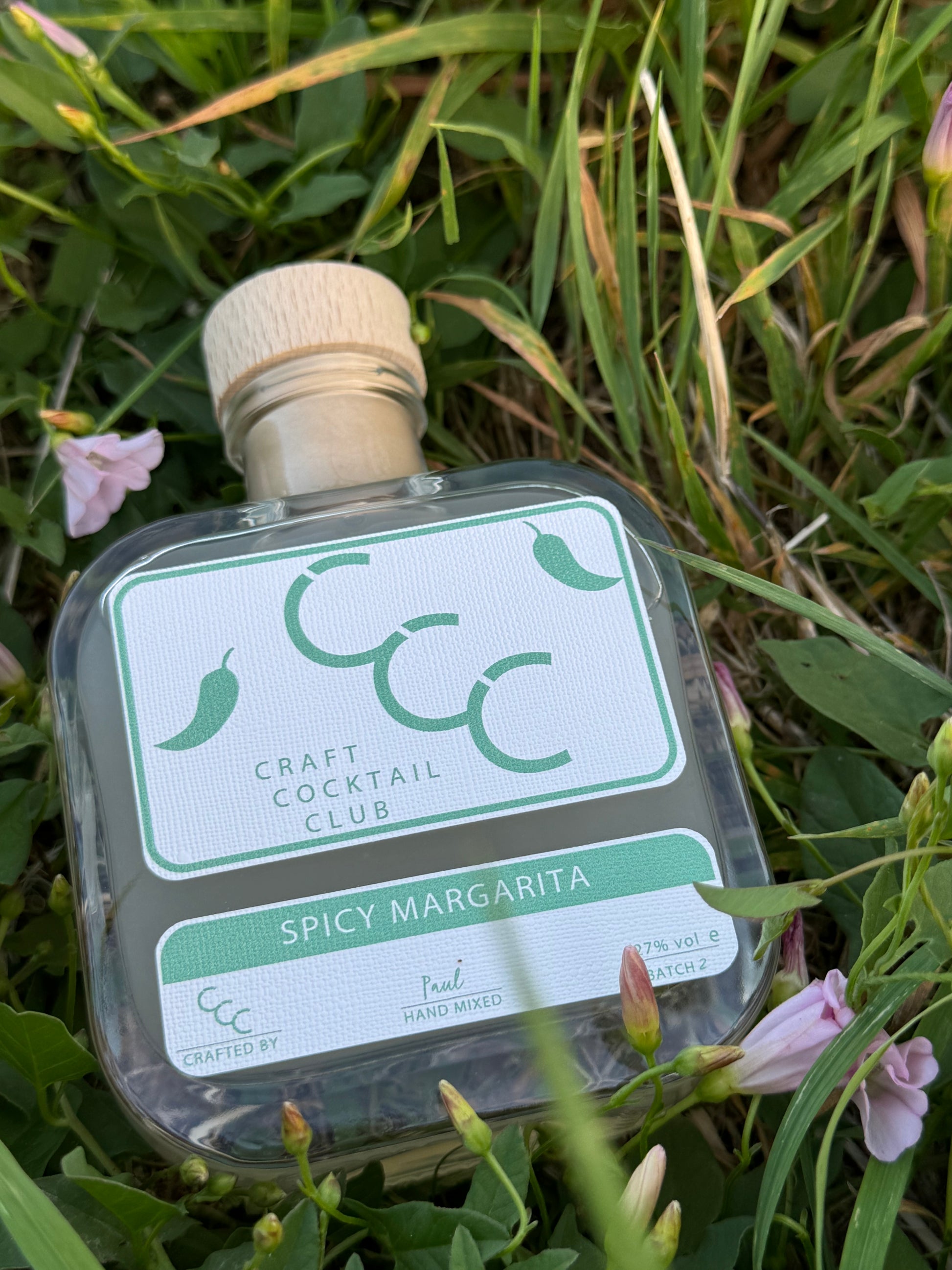 picture of a square cocktail bottle containing spicy margarita,  lying flat in some green grass and pink flowers 