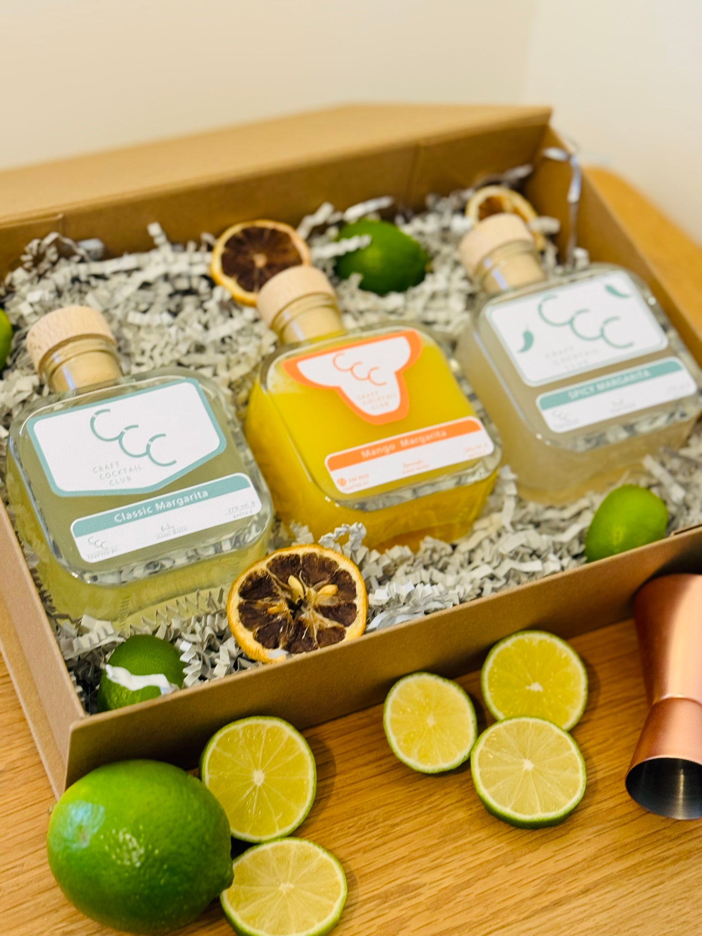 picture of a gift box with three margaritas  inside, the bottles a green and orange and. the box is surrounded by green fruit 