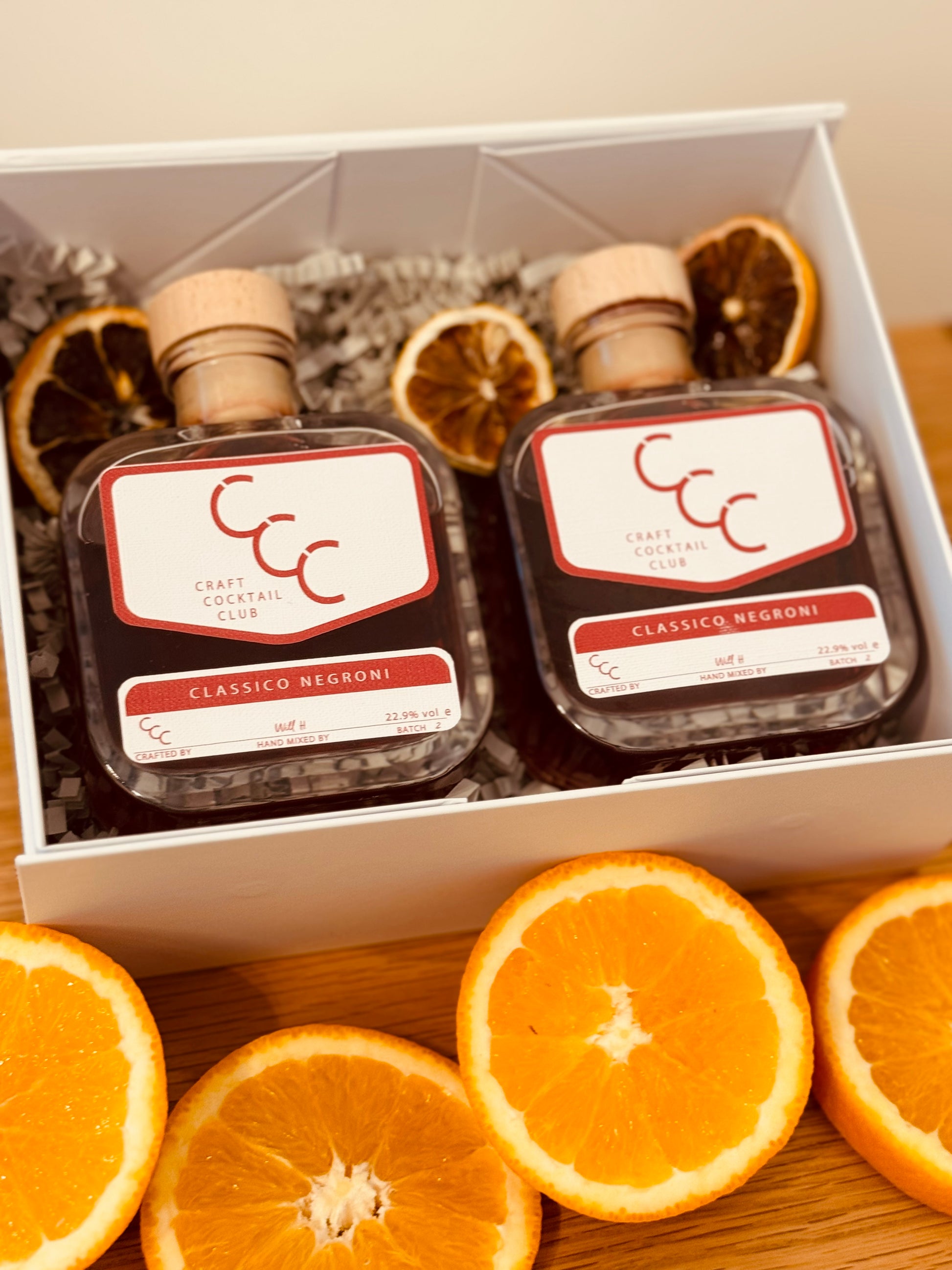 Picture of a gif box from the craft cocktail club, surrounded by freshly cut oranges. in the box are two glass square bottles that are full of dark red negroni 