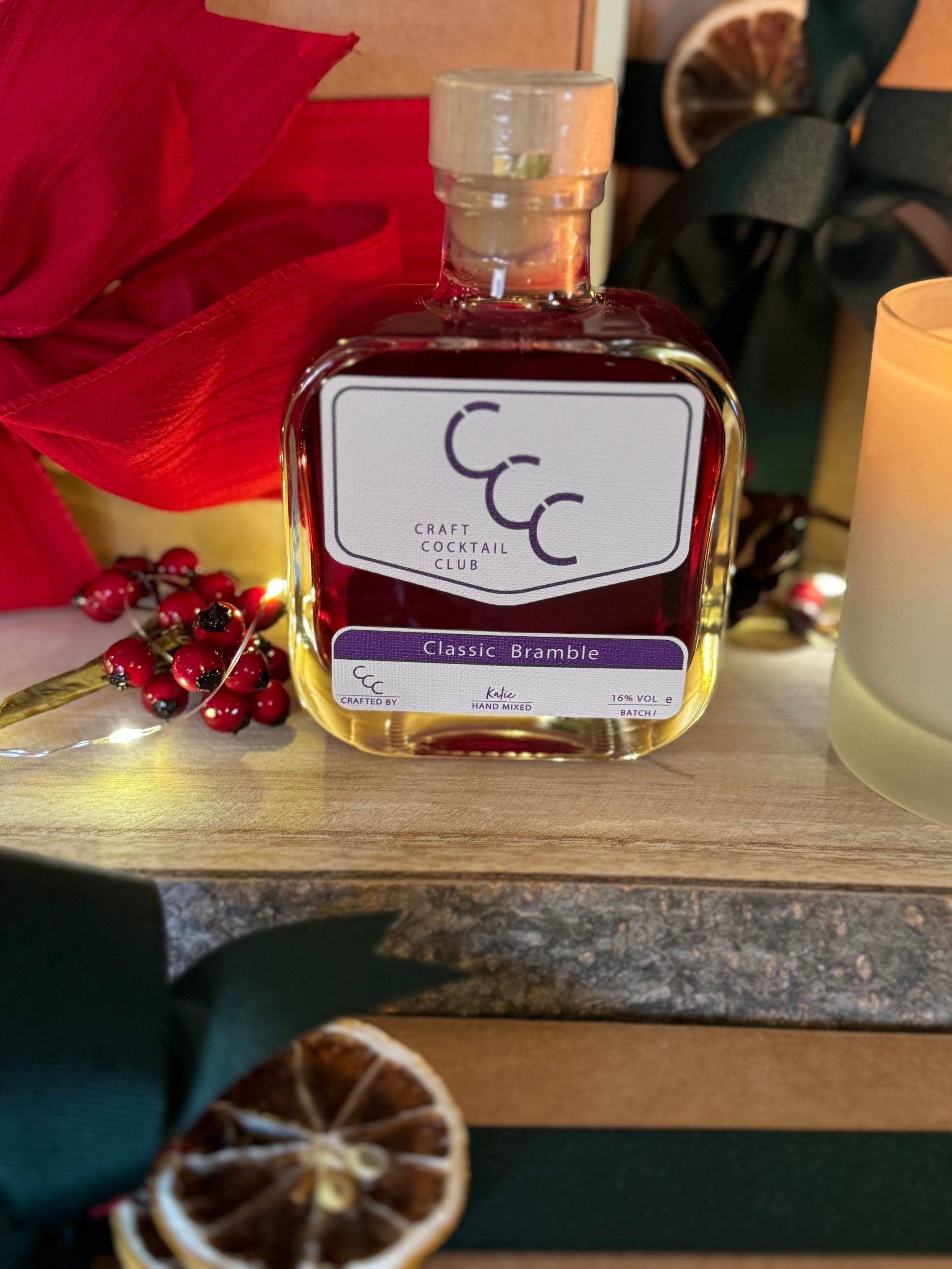 Picture  of a bottle of craft cocktail club pre mixed premium classic bramble cocktail, the liquid is deep purple and is surrounded by fair lights and gift boxes 