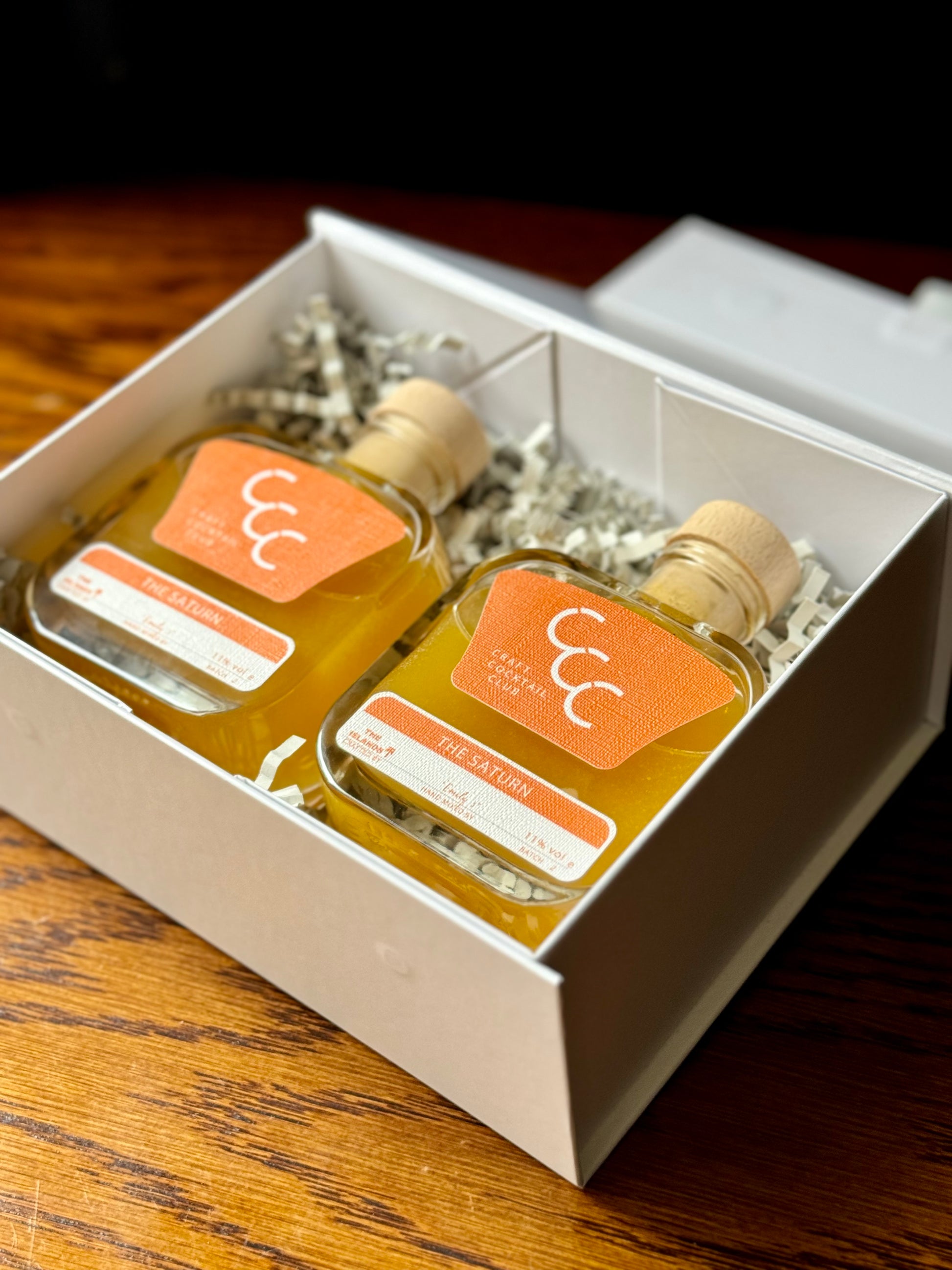 picture of pair of ready to drink orange cocktails in glass bottles in a white gift box on a wooden table 