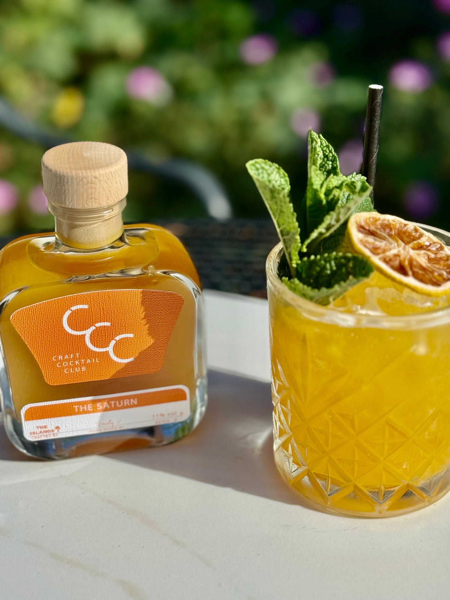 a sunny day with an orange cocktail with bit and orange slice on top next to a flower bed 