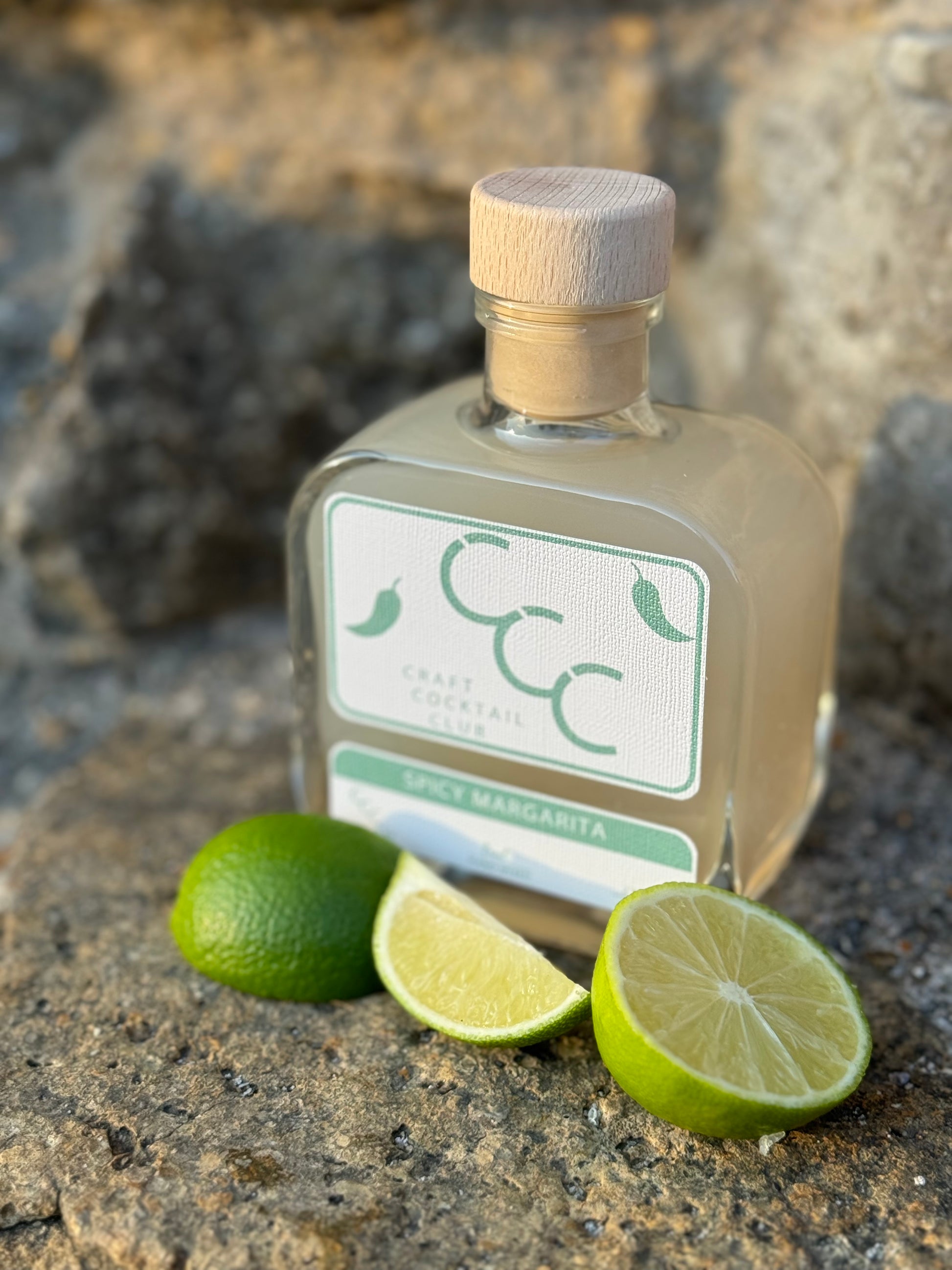 Picture of a square bottle on a limestone surface bottle contains craft cocktail spicy margarita and is surrounded by fresh cut limes 