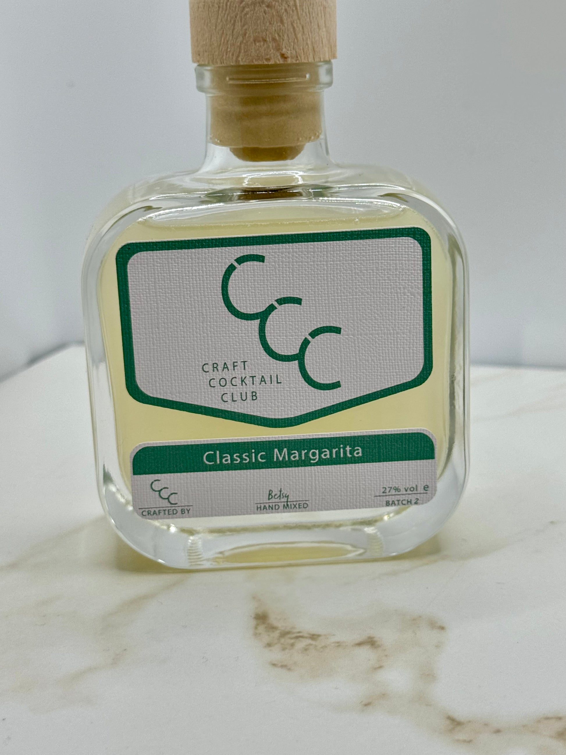 Picture of a small Square thick glass bottle with a cork, liquid inside is a lime green,  with a label that says craft cocktail club. 