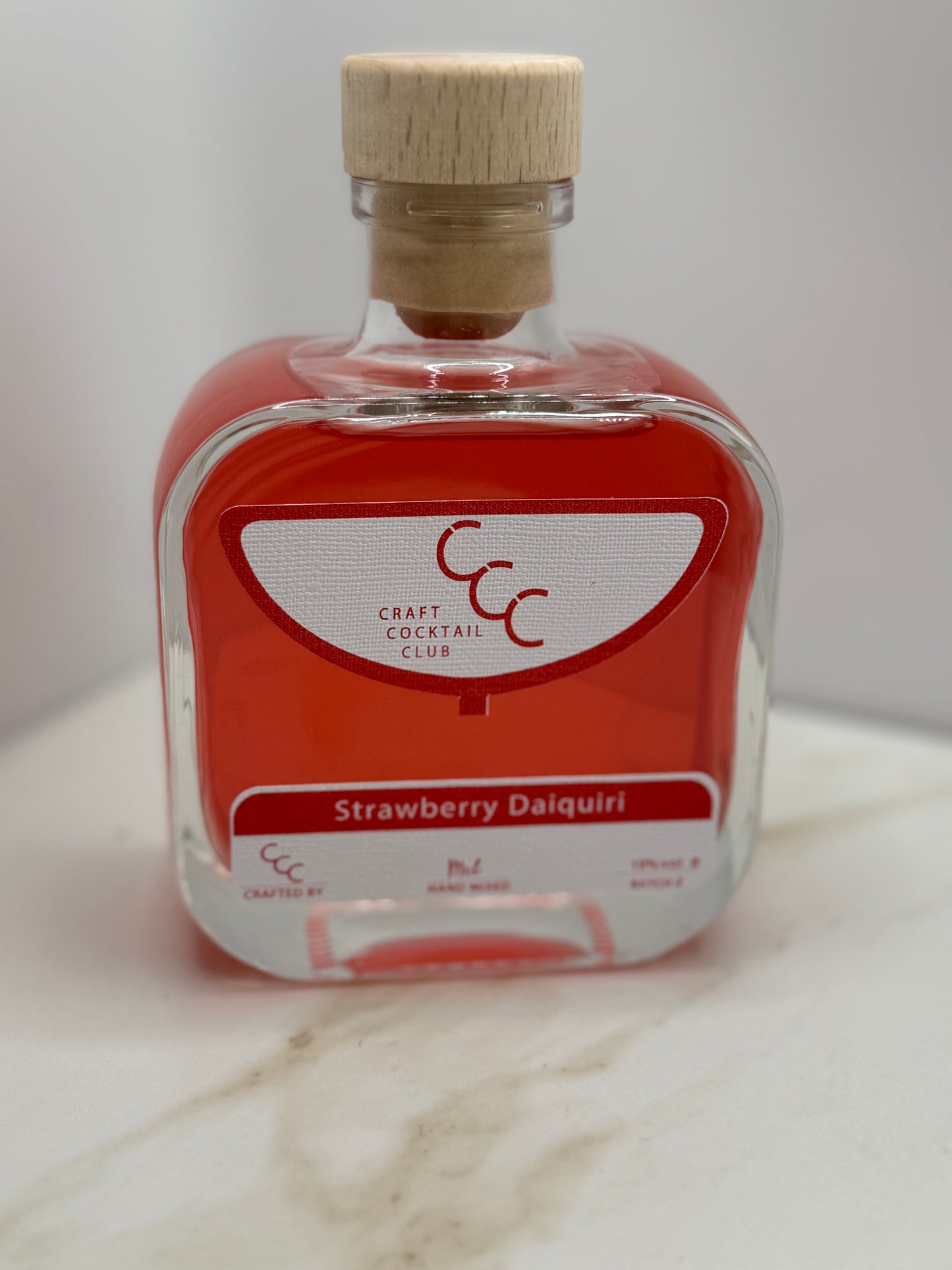 Picture of a Glass bottle that is square and made of thick glass, it has a wooden cork and a light red liquid inside. the label reads Strawberry Daiquiri   Craft Cocktail Club 