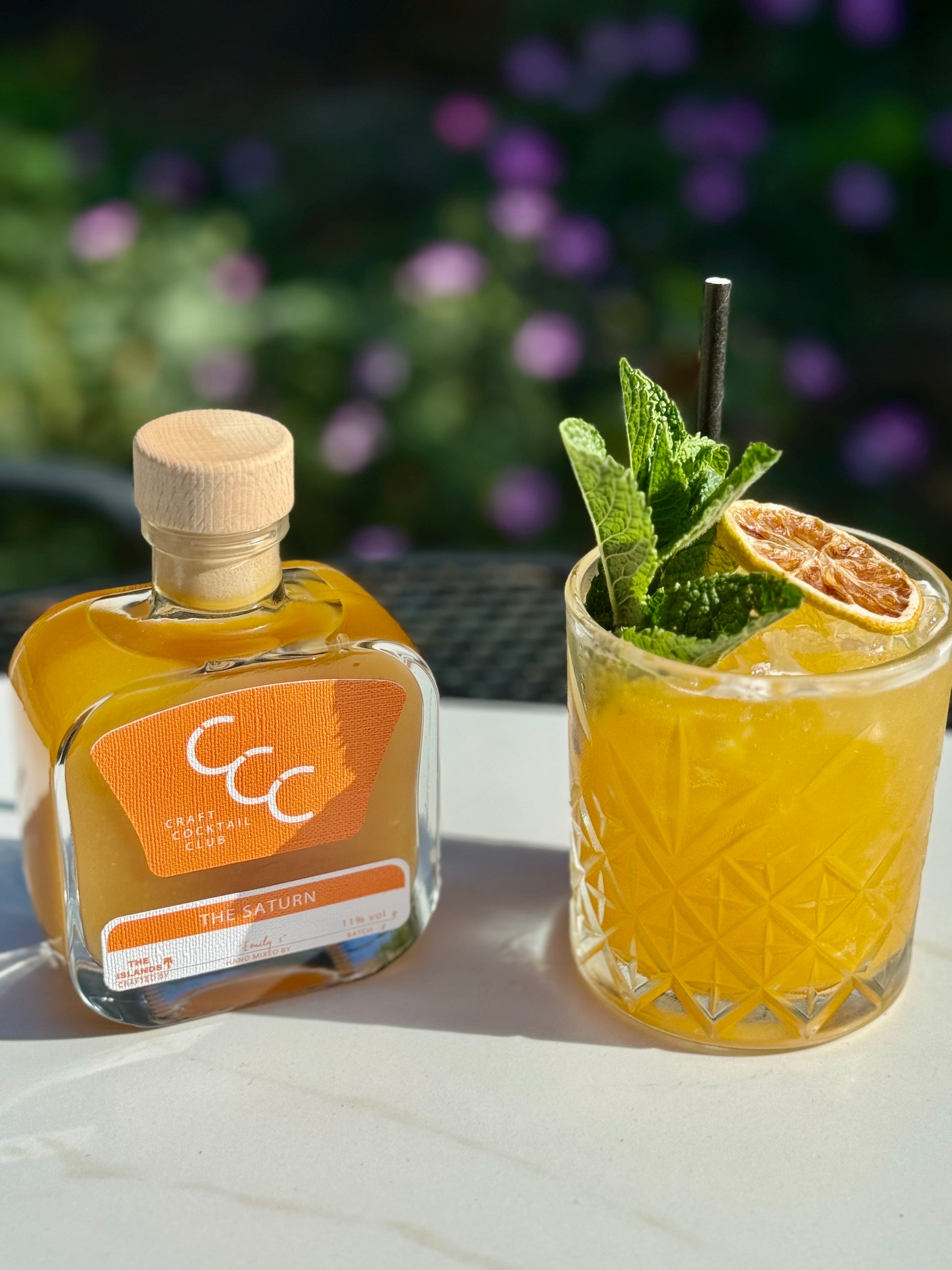 Picture  of a Saturn cocktail next to a square glass bottle, set outside on a sunny day with purple flowers in the background 