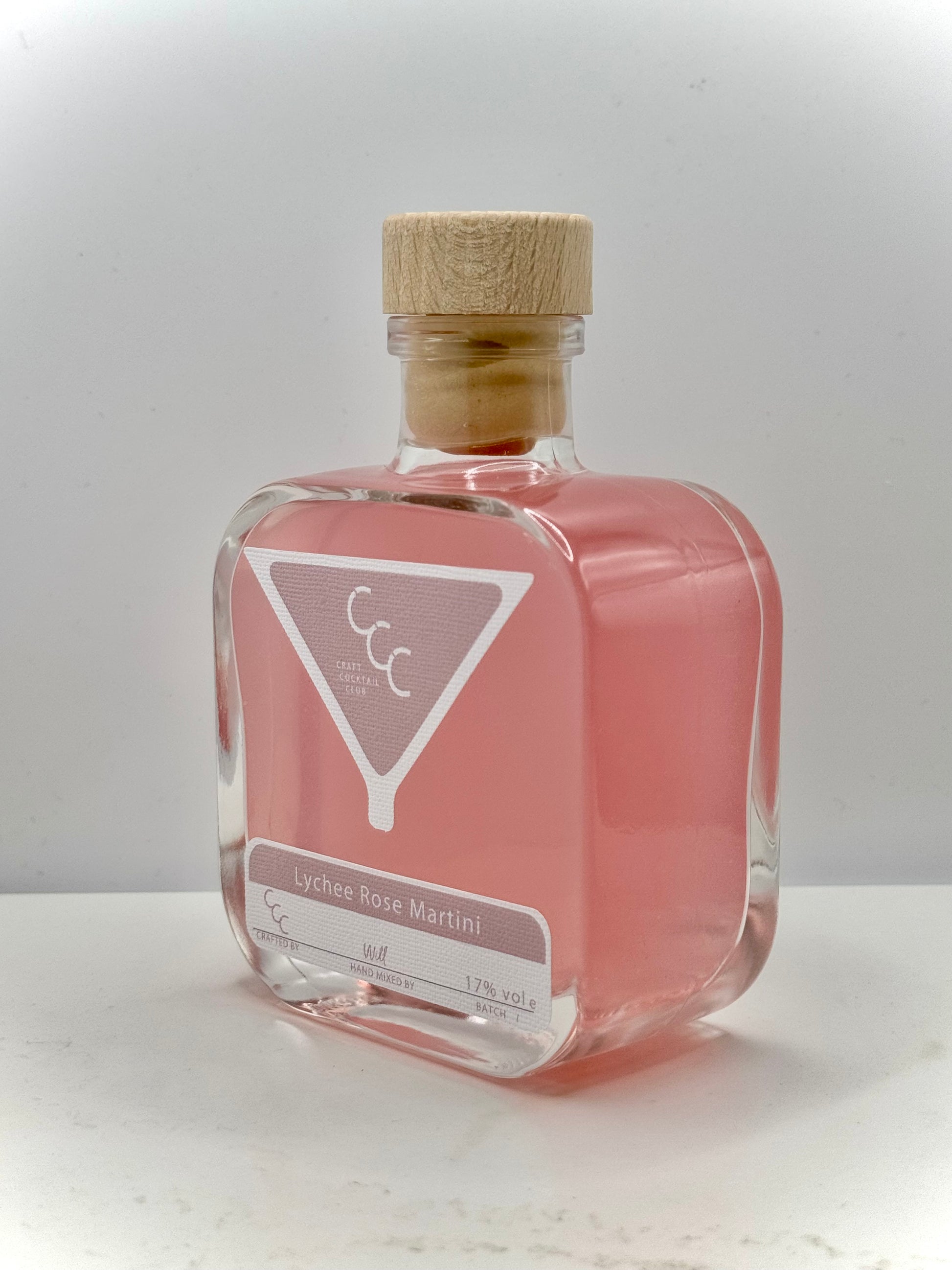 Picture of a small Square thick glass bottle with a cork, liquid inside is a light pink with a label that says craft cocktail club