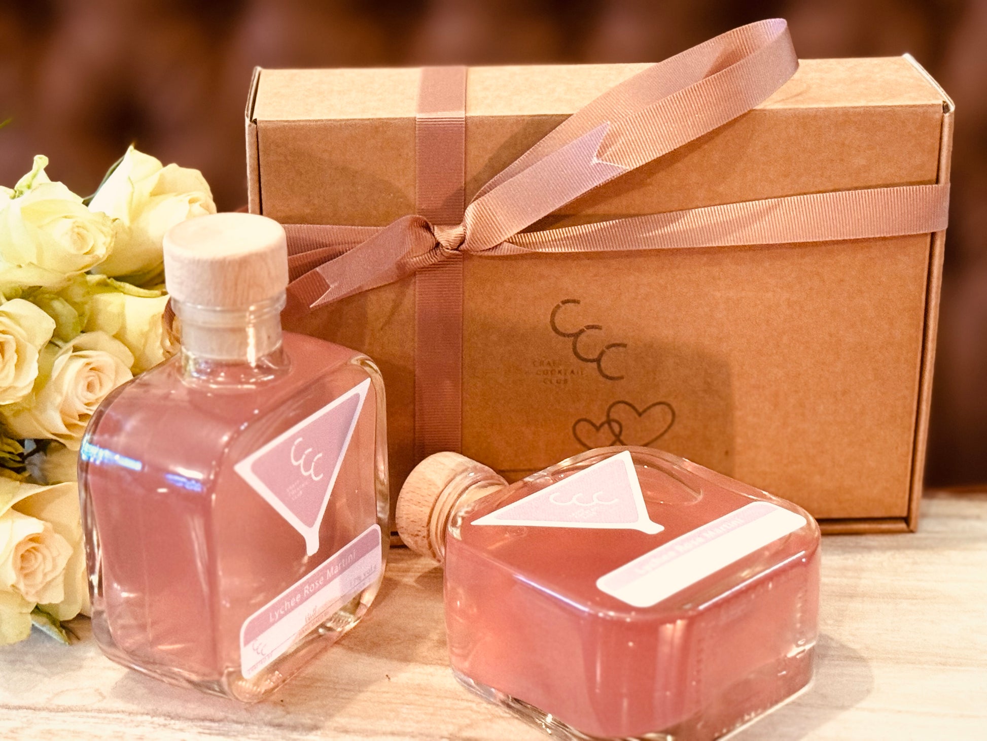 Picture of two pink bottles of Cocktails next to a gift box with white roses in the background 