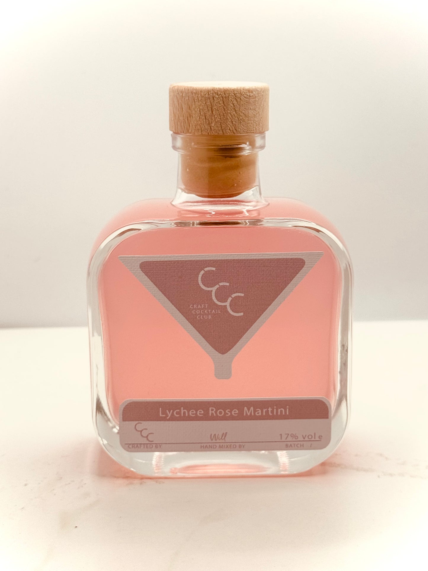 Picture of a pink Square glass bottles of Cocktail, on a marble surface.  Label reads Craft Cocktail Club. Lychee rose martini 