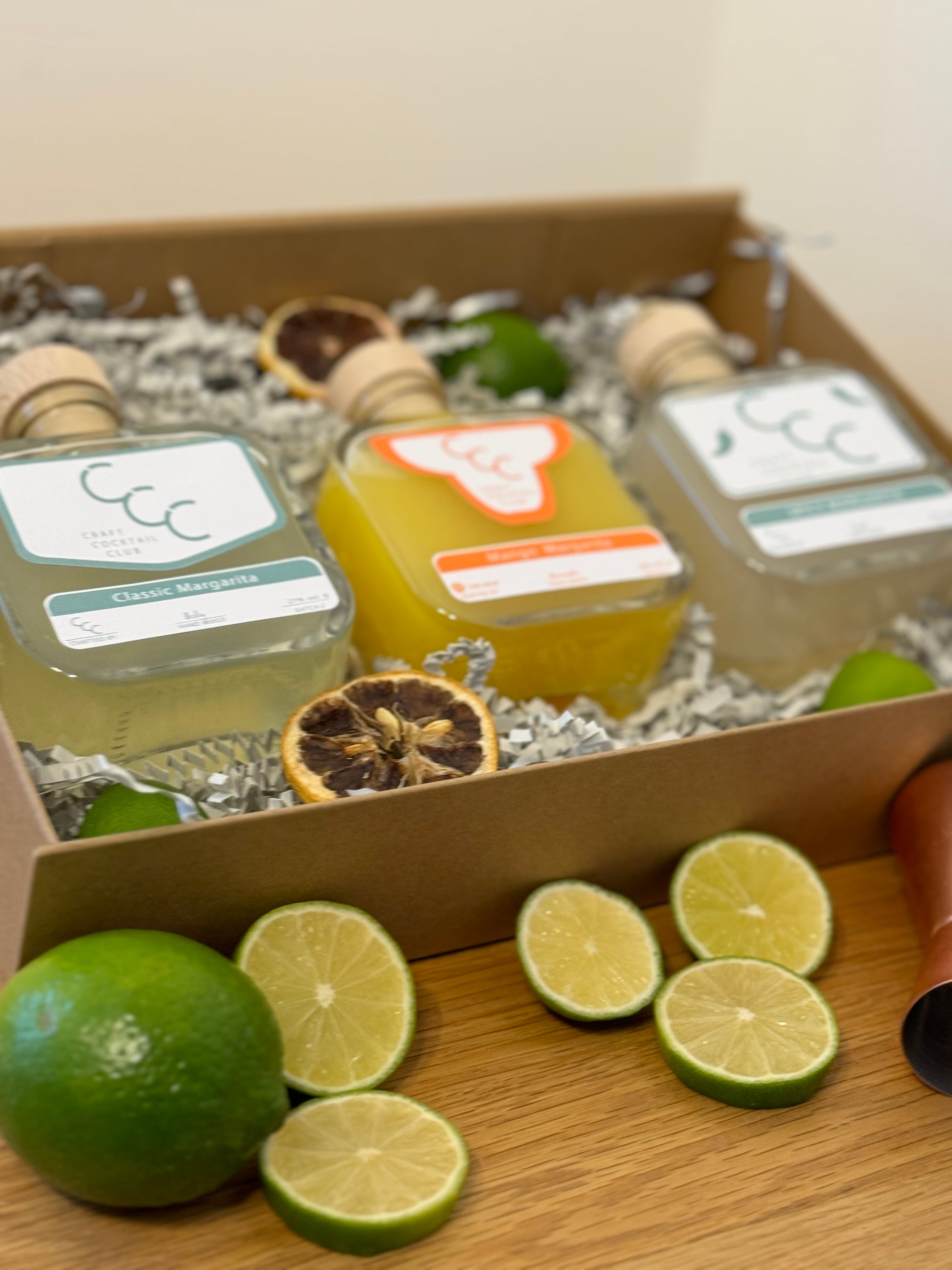 picture of a gift box with three margaritas  inside, the bottles a green and orange and. the box is surrounded by green fruit 