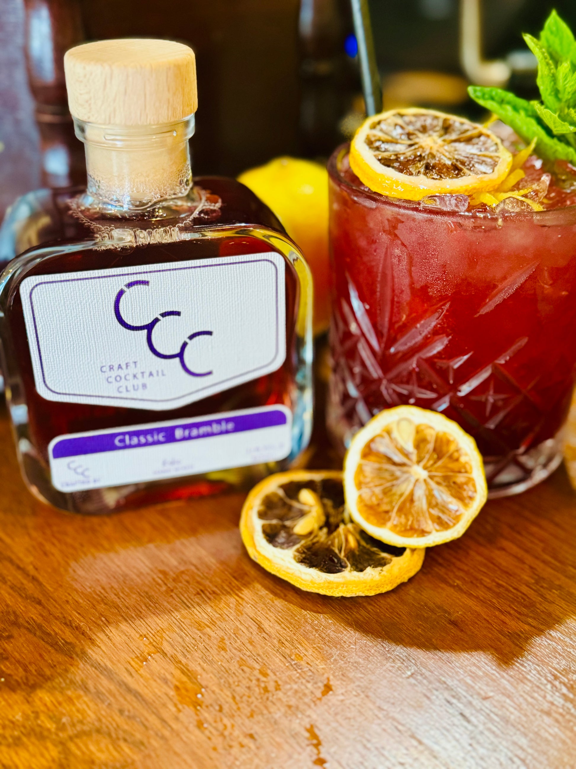 picture of a bramble gin cocktail next to a bottle of craft cocktail club and lemons 