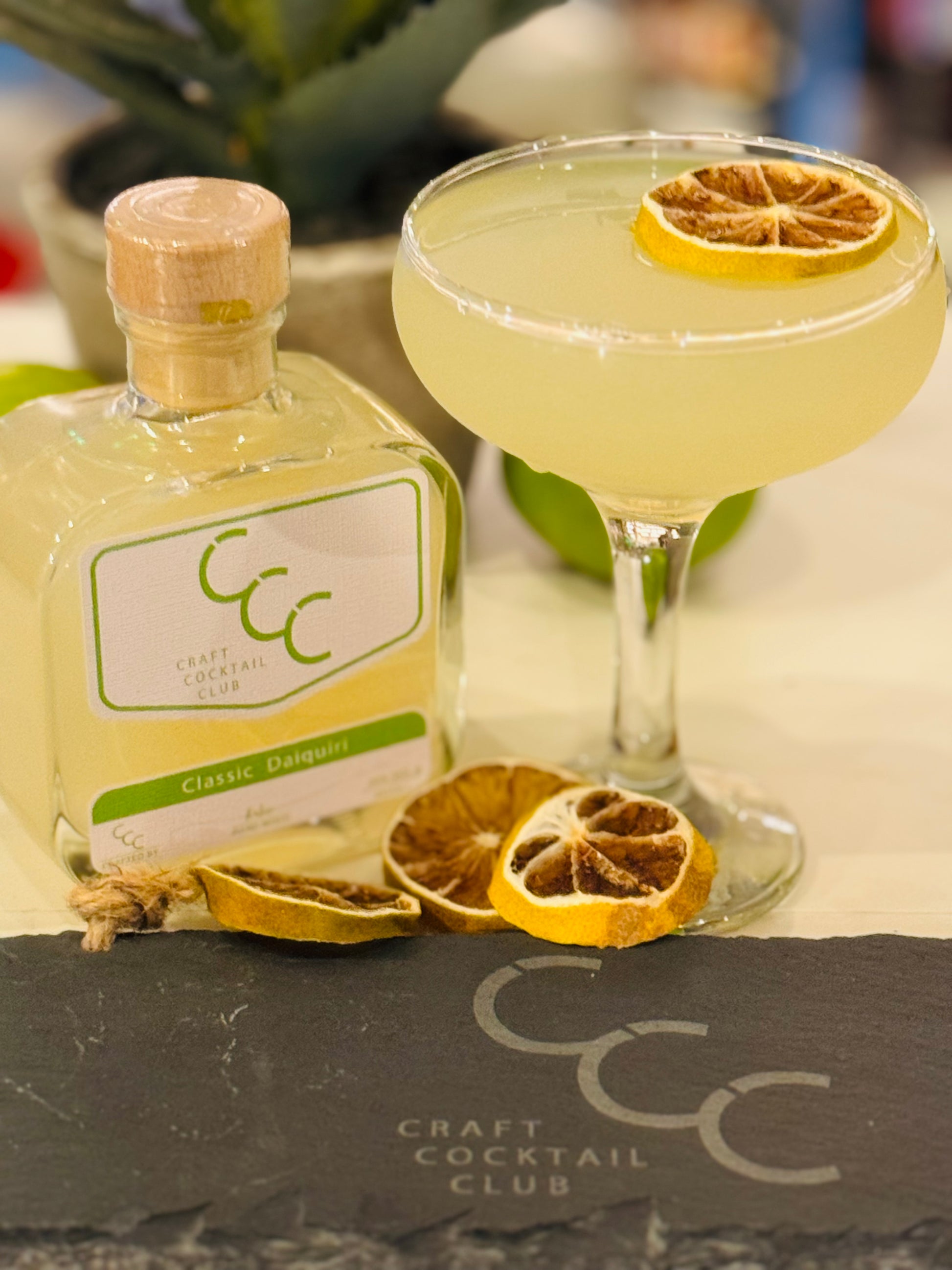 Picture of a Craft Cocktail Club Bottle on a bar sat next to a coup glass full of lime coloured liquid 