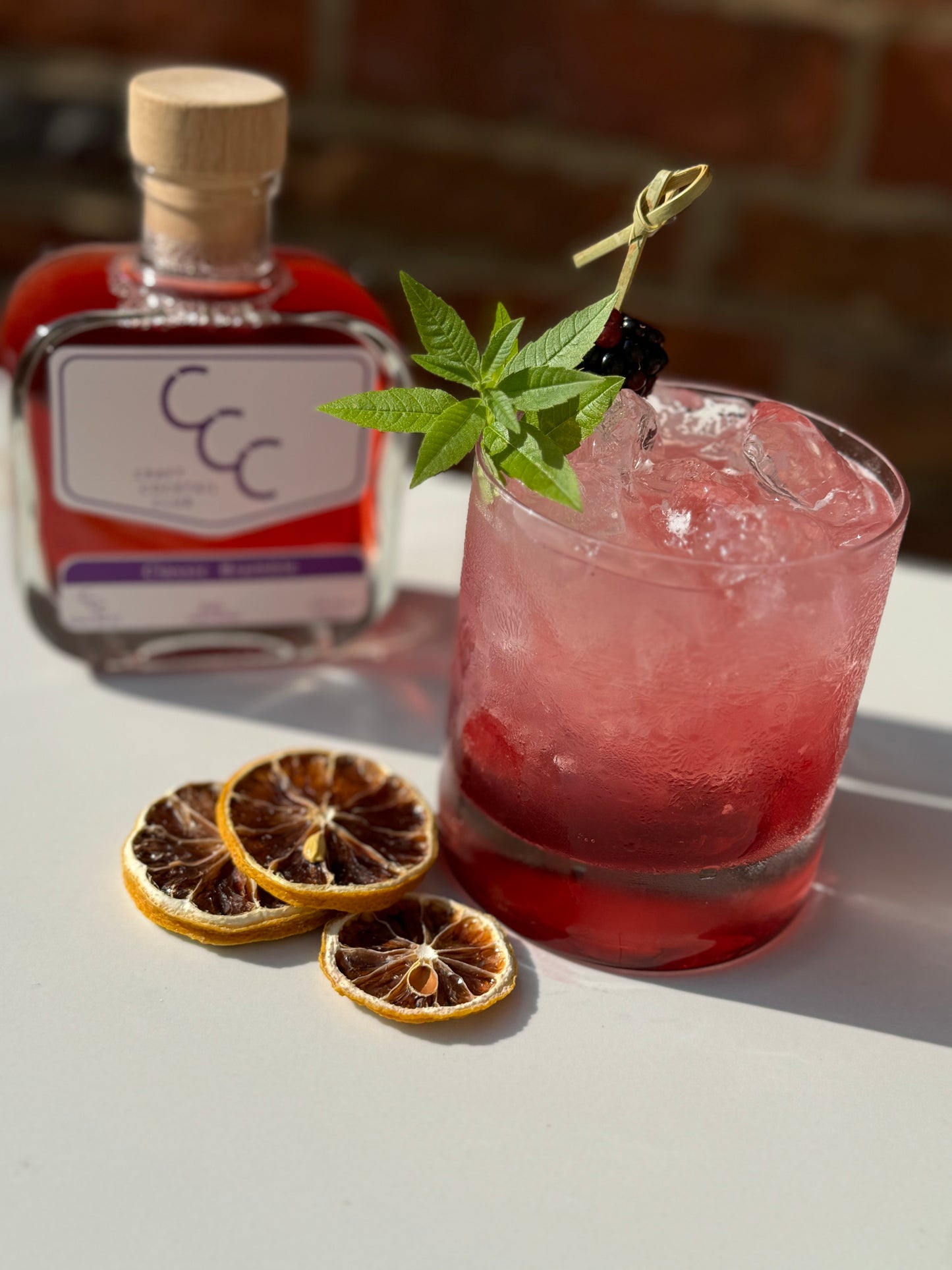 Picture of a craft cocktail square bottle next to a purple glass filled. with bramble cocktail with a mint garnish 