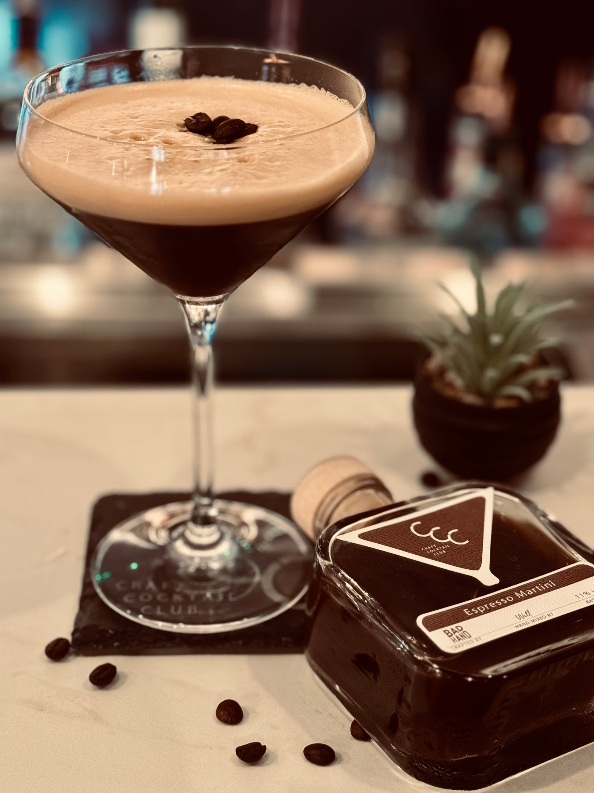 Picture of a Cocktail in a martini glass, with a square bottle of ready to drink cocktail next to it on a whit  tile with coffee bean around it, label reads craft cocktail club espresso martini