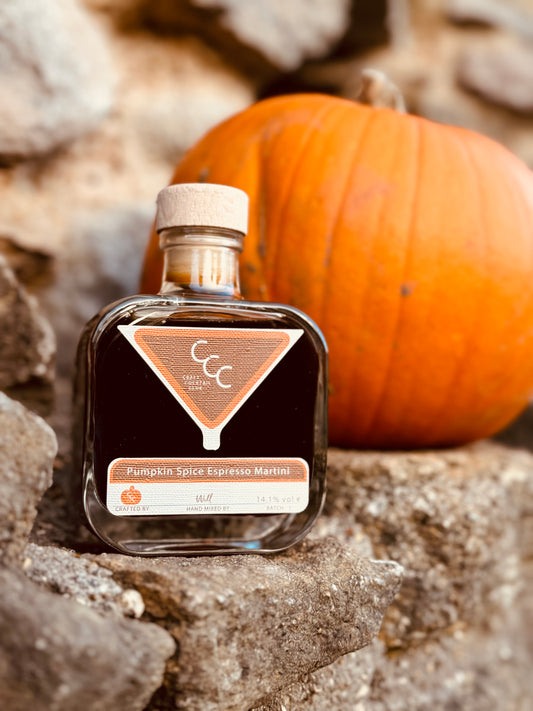 Picture of a square glass bottle with a dark brown liquid, label reads pumpkin spice espresso martini, picture is taken on a sandstone rock with a pumpkin in the background 