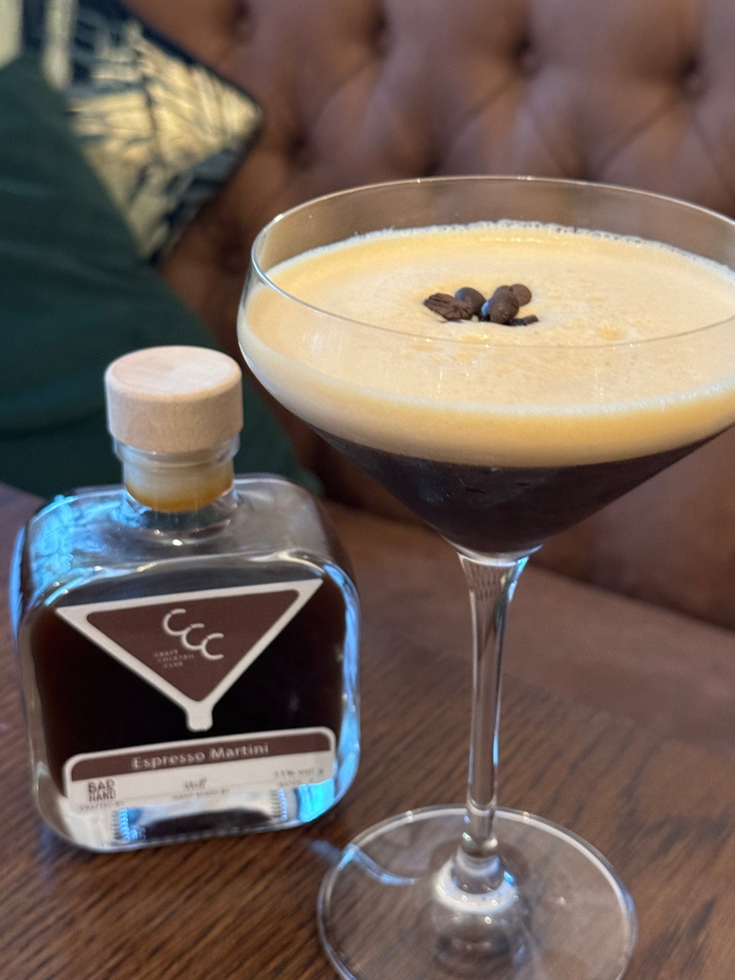 Picture of a cocktail in a martini glass, liquid is a dark brown with a light foam with three coffee beans on top, sat on a dark wood table with cushions in the background 