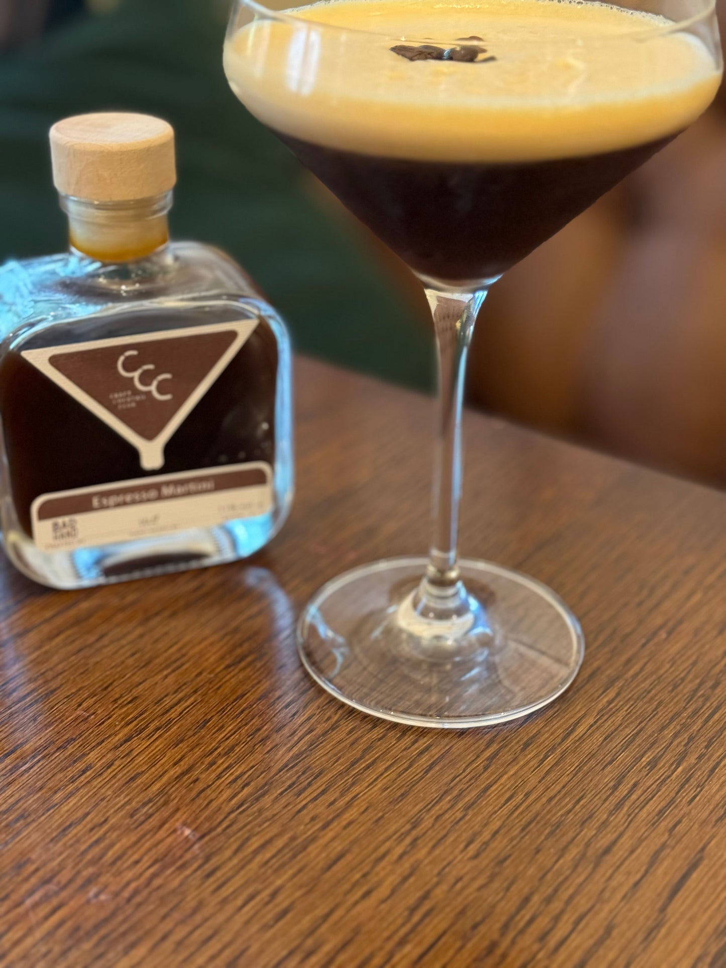 Picture of an espresso martini in a glass with three coffee beans on top, sat on a dar wood table with a craft cocktail club bottle next to it 