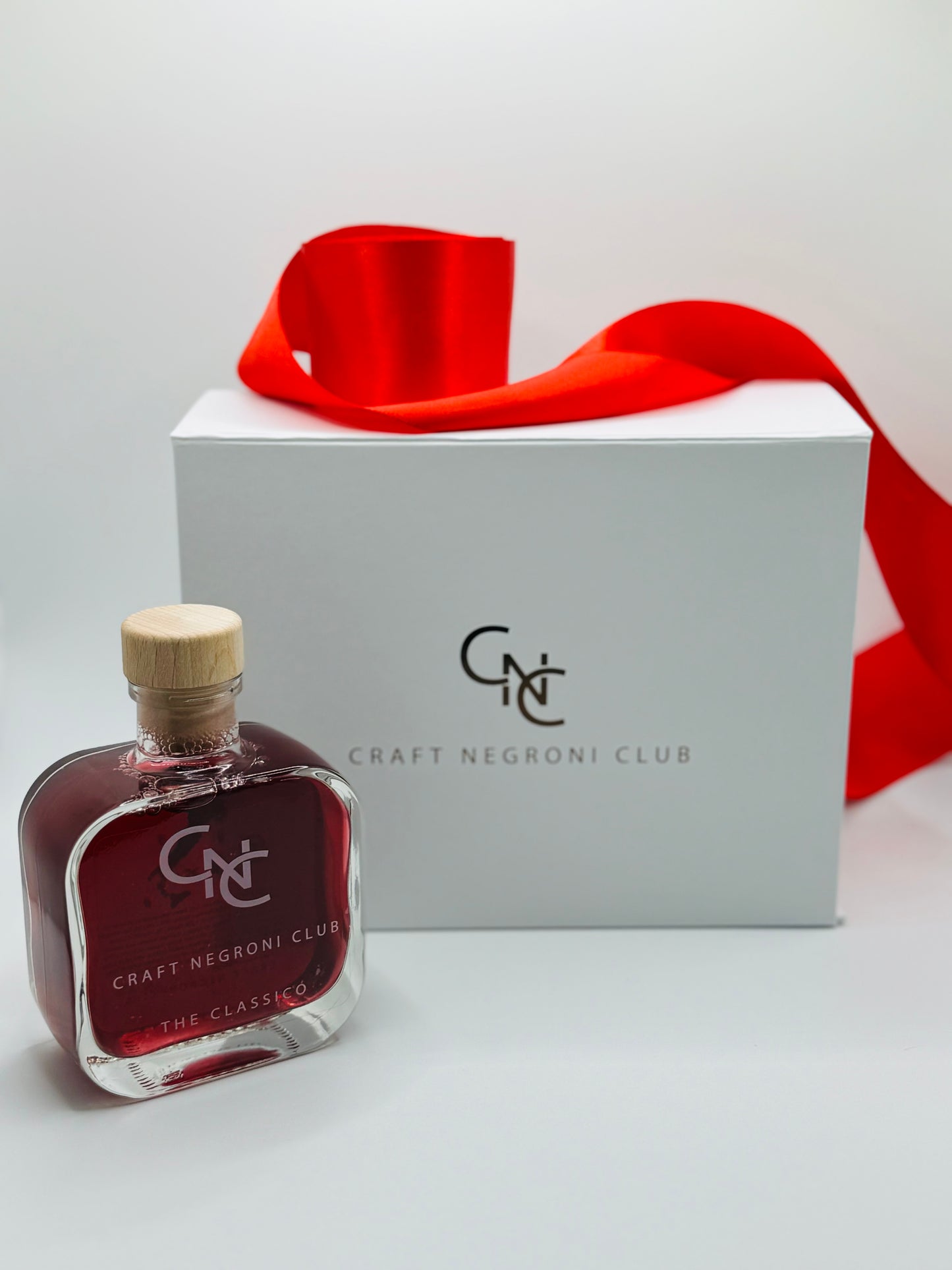 picture of a Craft Negroni Club gift box with a red ribbon on top, with a single square bottle of red liquid next it on the left 