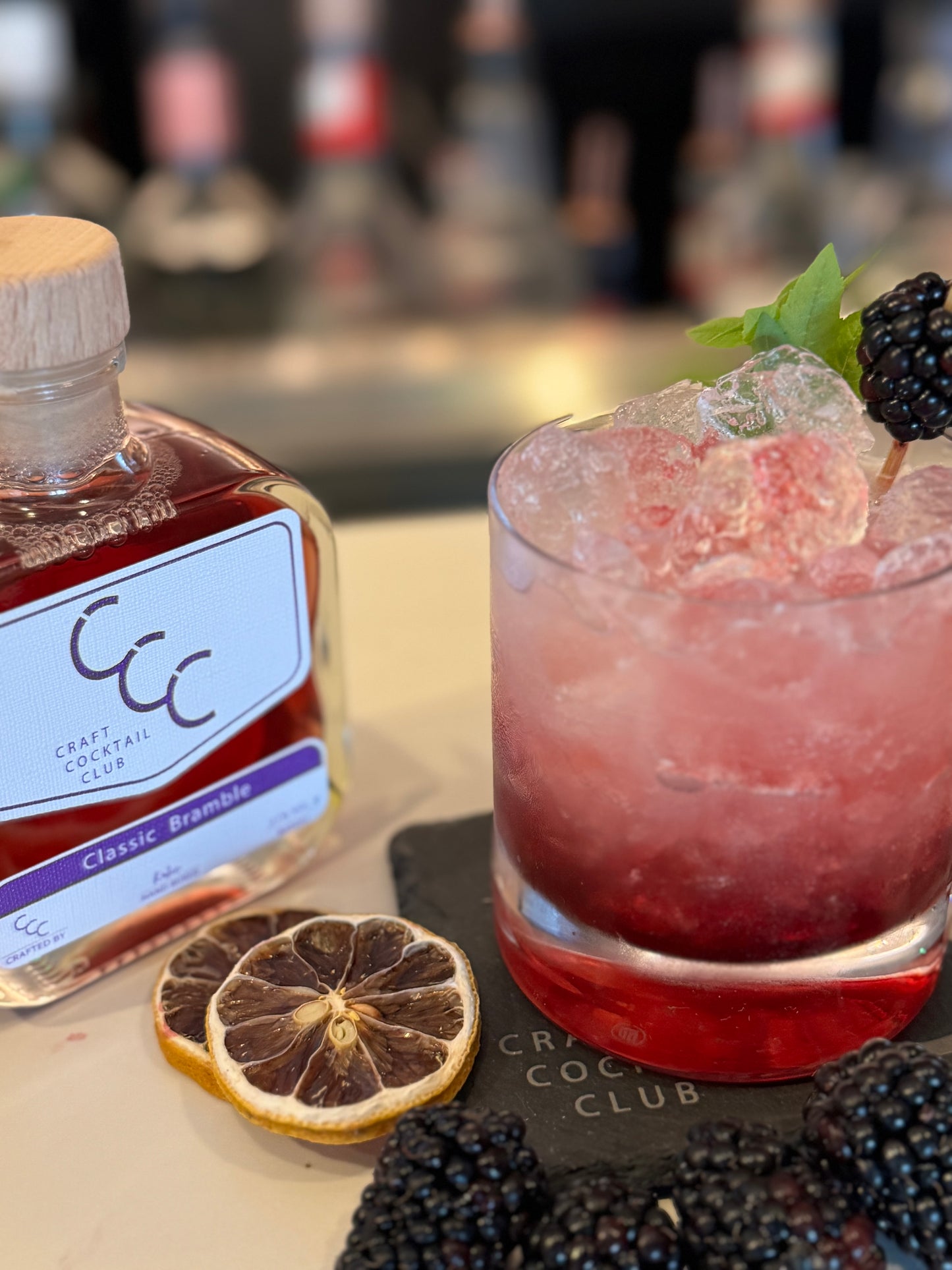 picture of a bramble cocktail surrounded by fruit, on a slate coaster in bar next to a craft cocktail club bottle 