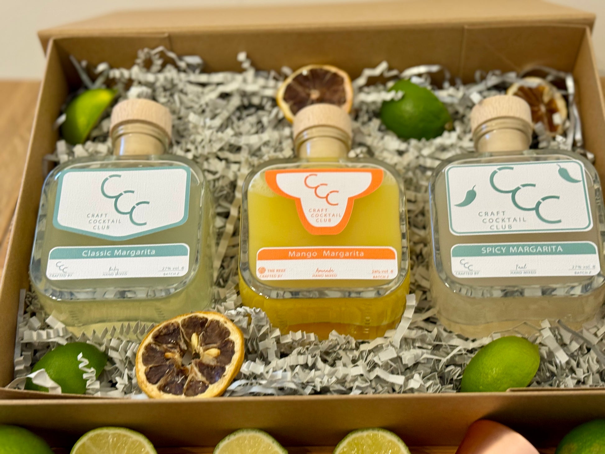 picture of a gift box with three margaritas  inside, the bottles a green and orange and. the box is surrounded by green fruit 