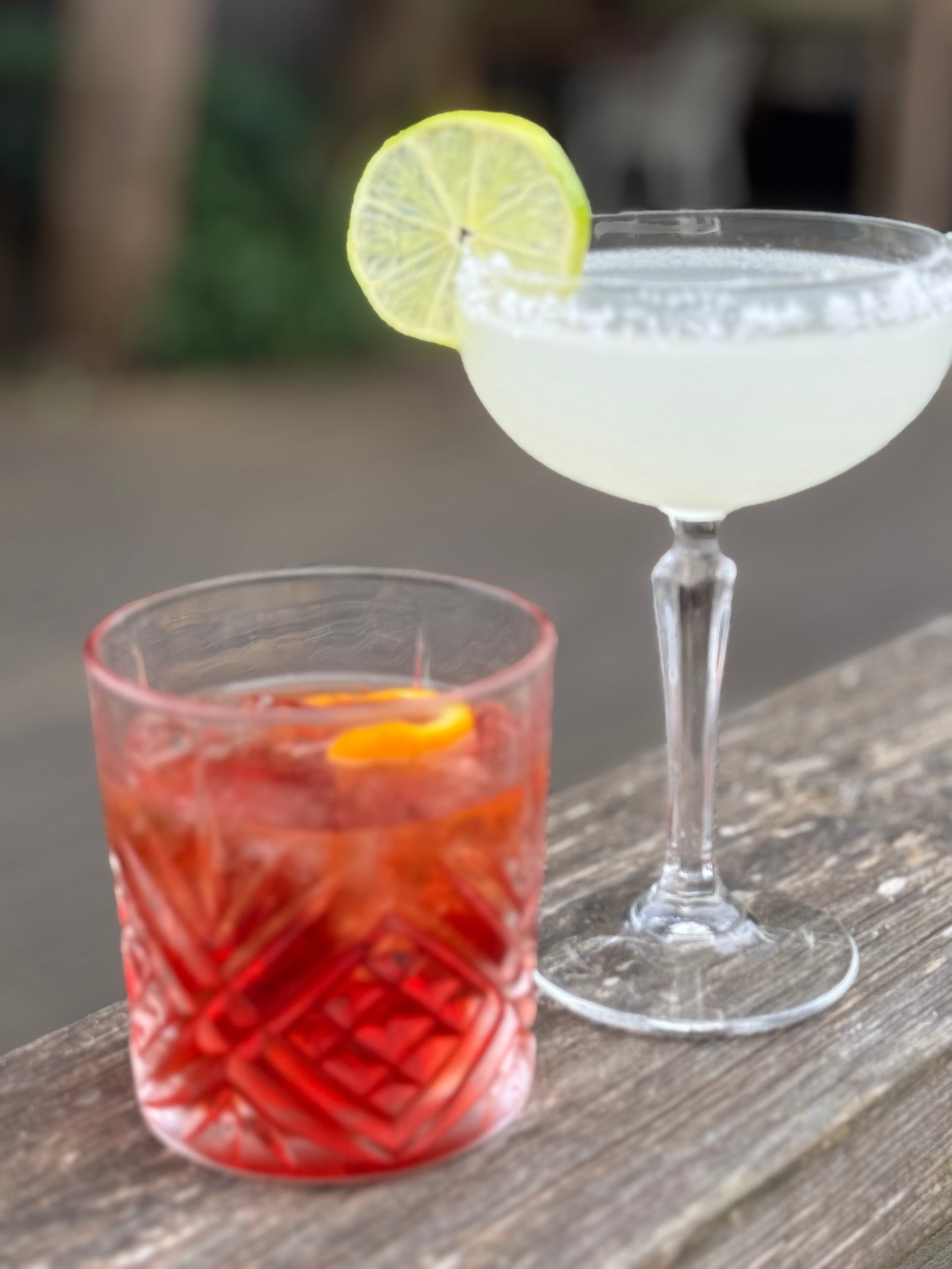 picture o a margarita with a lime wheel next to a red negroni glass outside on a bench