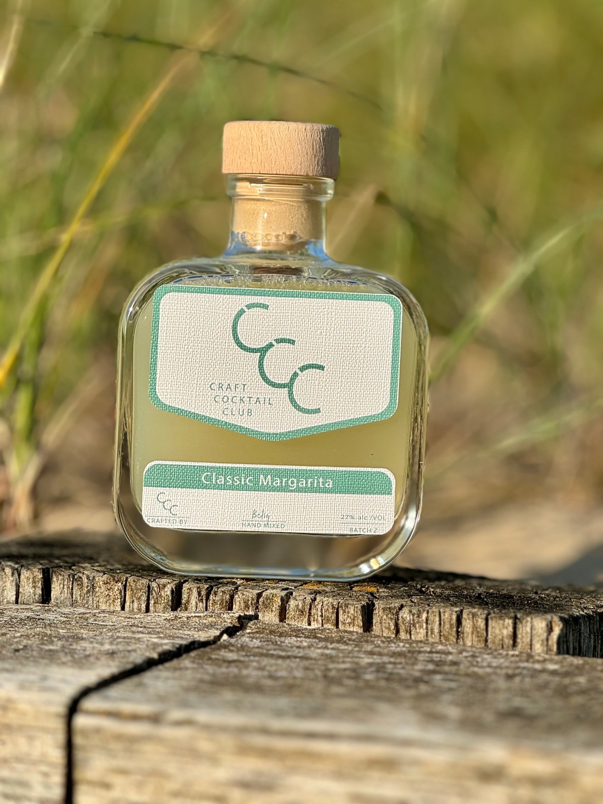 picture of a ready made margarita cocktail outside on a drift wood pile with beach and grass in the background 