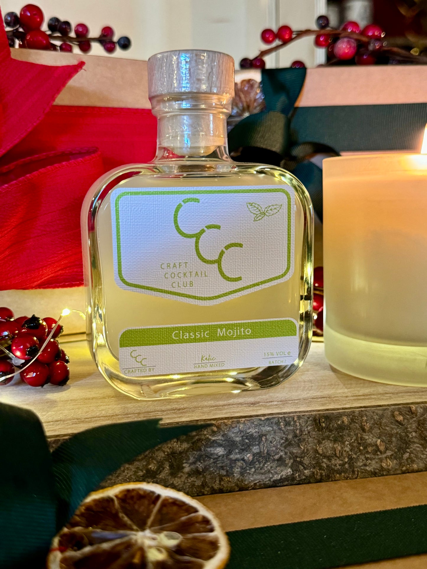 picture of a bottle of mojito next to a candle surround by christmas gifts with red and green ribbon 