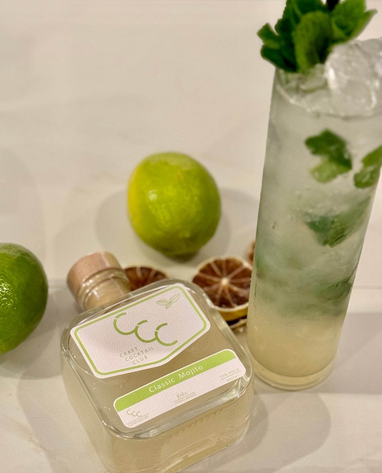 Picture of a craft cocktail club square glass bottle next to a complete tall glass of mojito in a glass with lots of ice and fresh mint