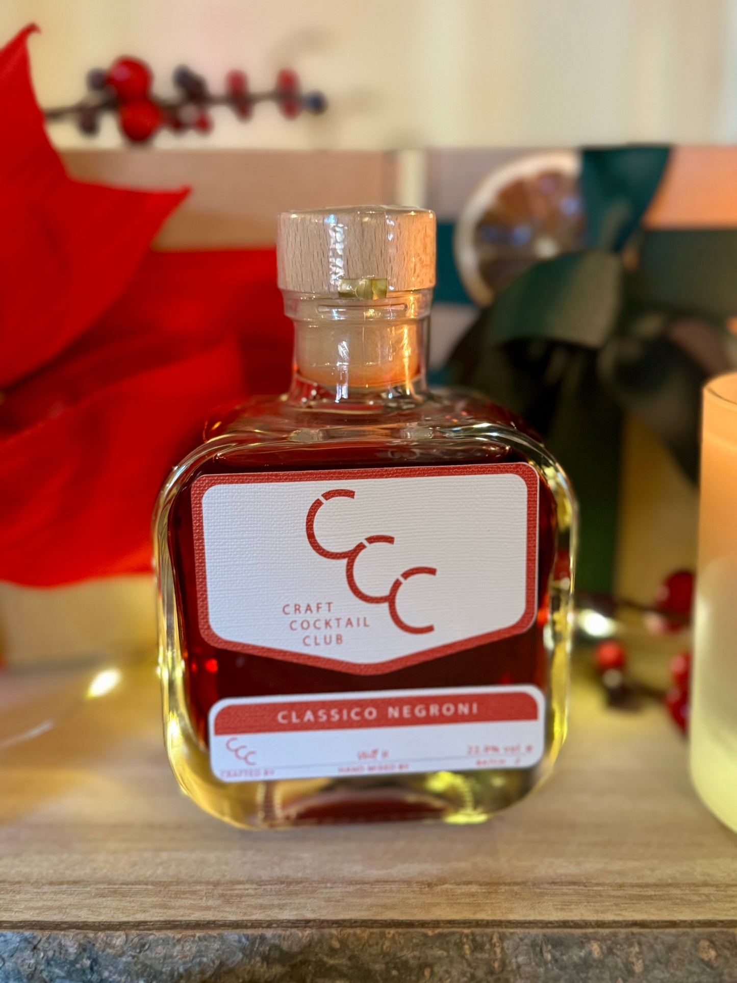 A picture of a craft cocktail club. bottle of ready made Christmas negroni, with fairly lights in the background with 2 gifs with green and red ribbon 