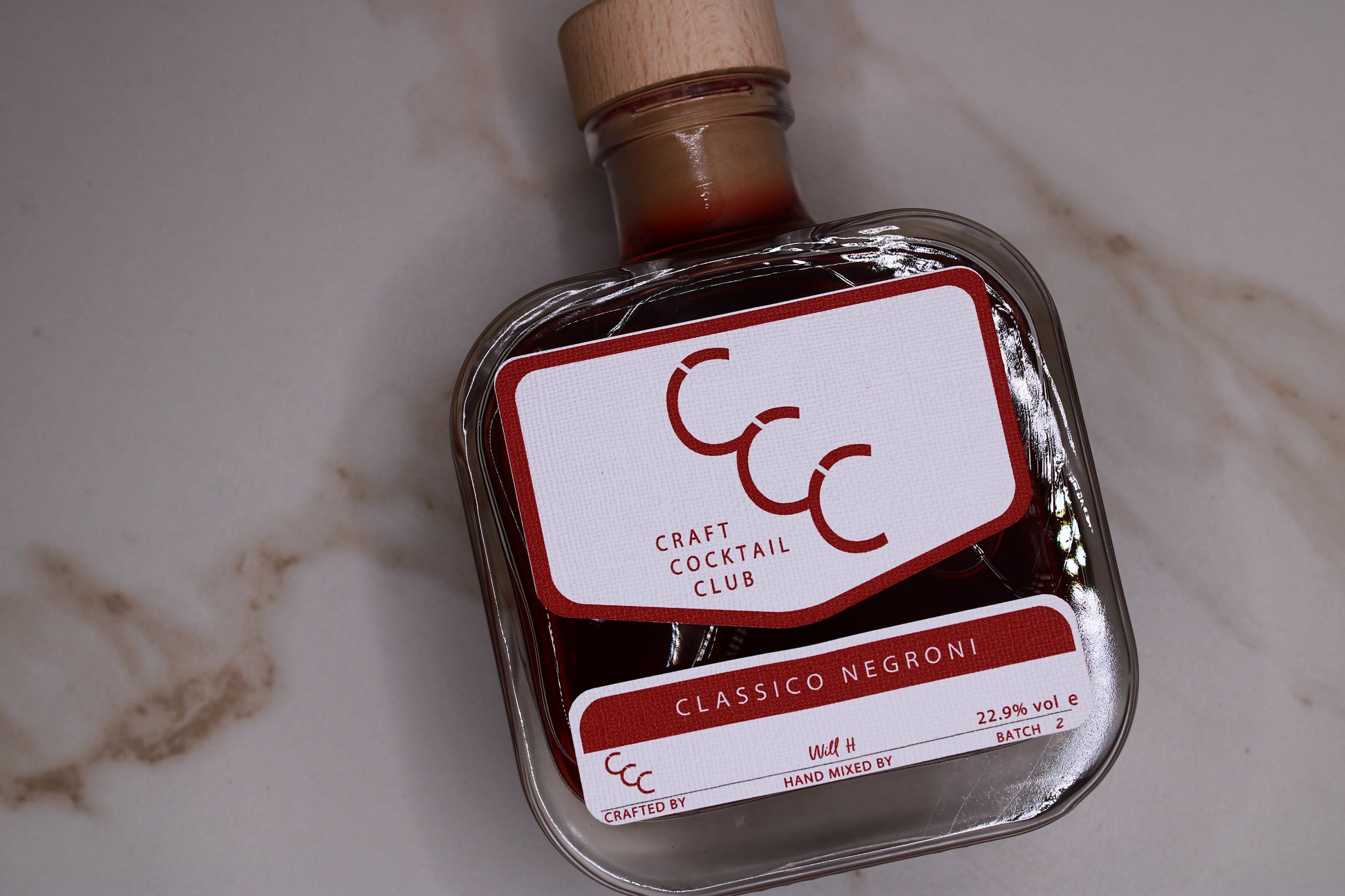 Picture  of a sign bottle of ready to drink negroni in a glass bottle, lying flay on a cream marble tile with a brown streak. label reads craft cocktail club, Classic Negroni Recipe 