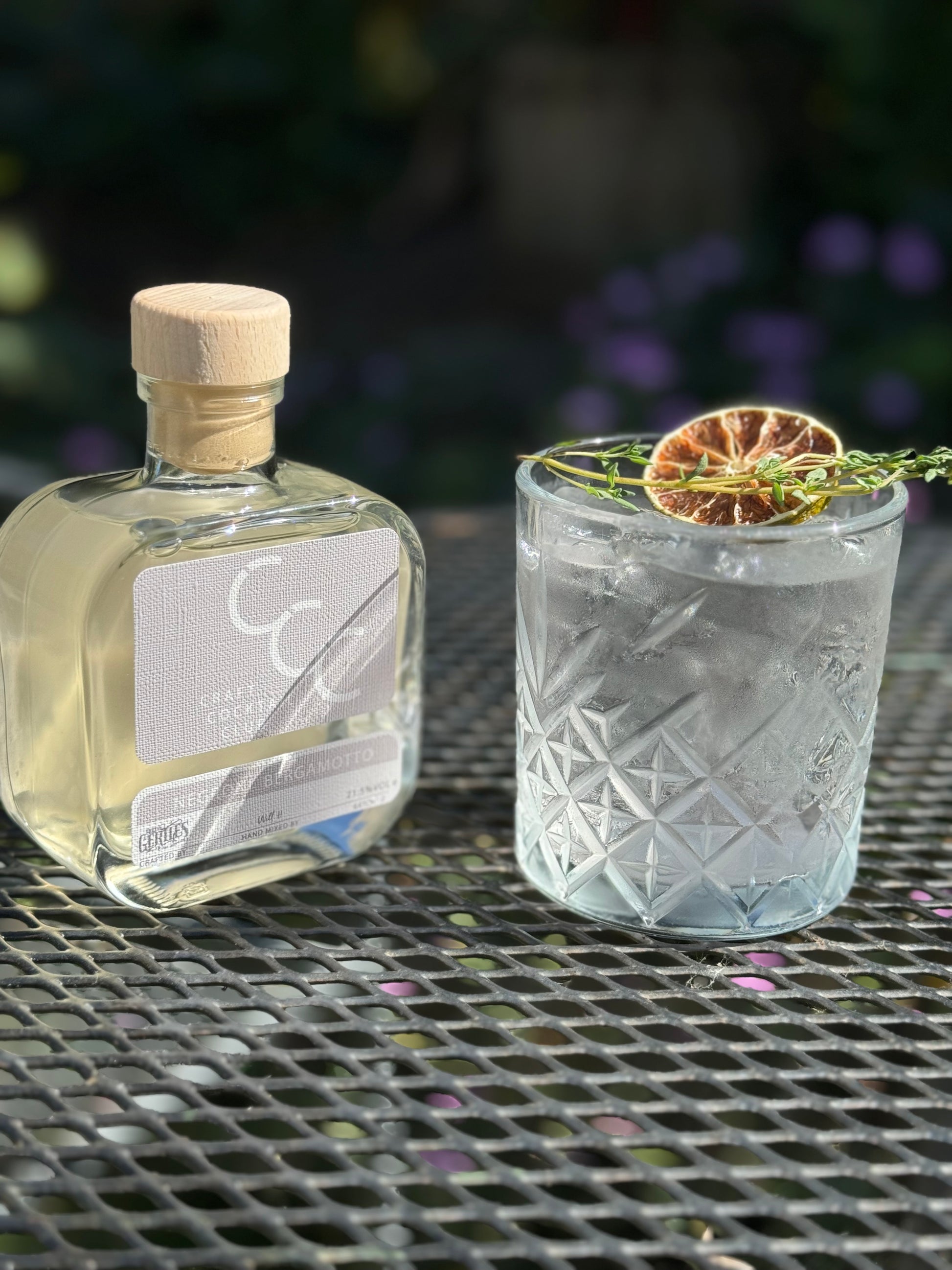 outdoor picture of a white negroni drink next to a bottle purple flowers blur in the background 