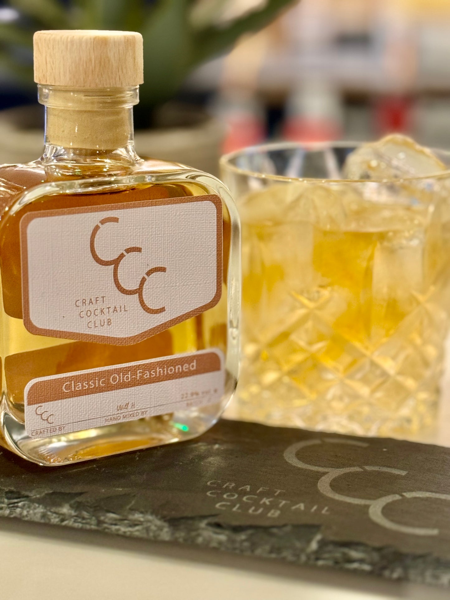 A Picture of  a small square thick glass bottle sat on a slate bar with a picture of an Old Fashioned Cocktail behind it, containing a golden liquid and ice. Label on the bottle says Craft Cocktail Club, Classic Old- Fashioned Cocktail Ready to drink