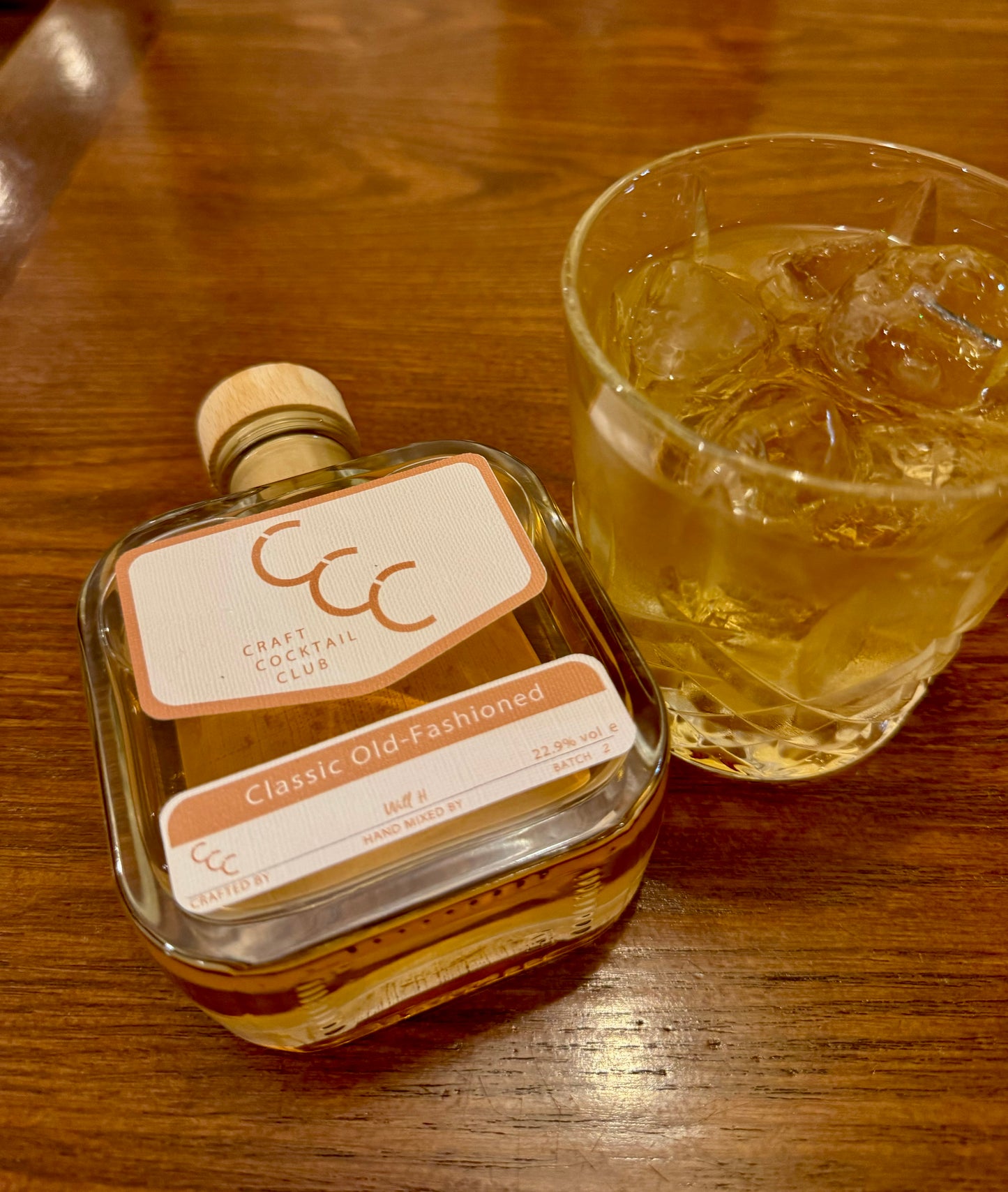 Picture of a Ready to drink old fashioned cocktail bottle lying flat on a wood table., next to it is a crystal glass full of a golden cocktail and loads of ice 