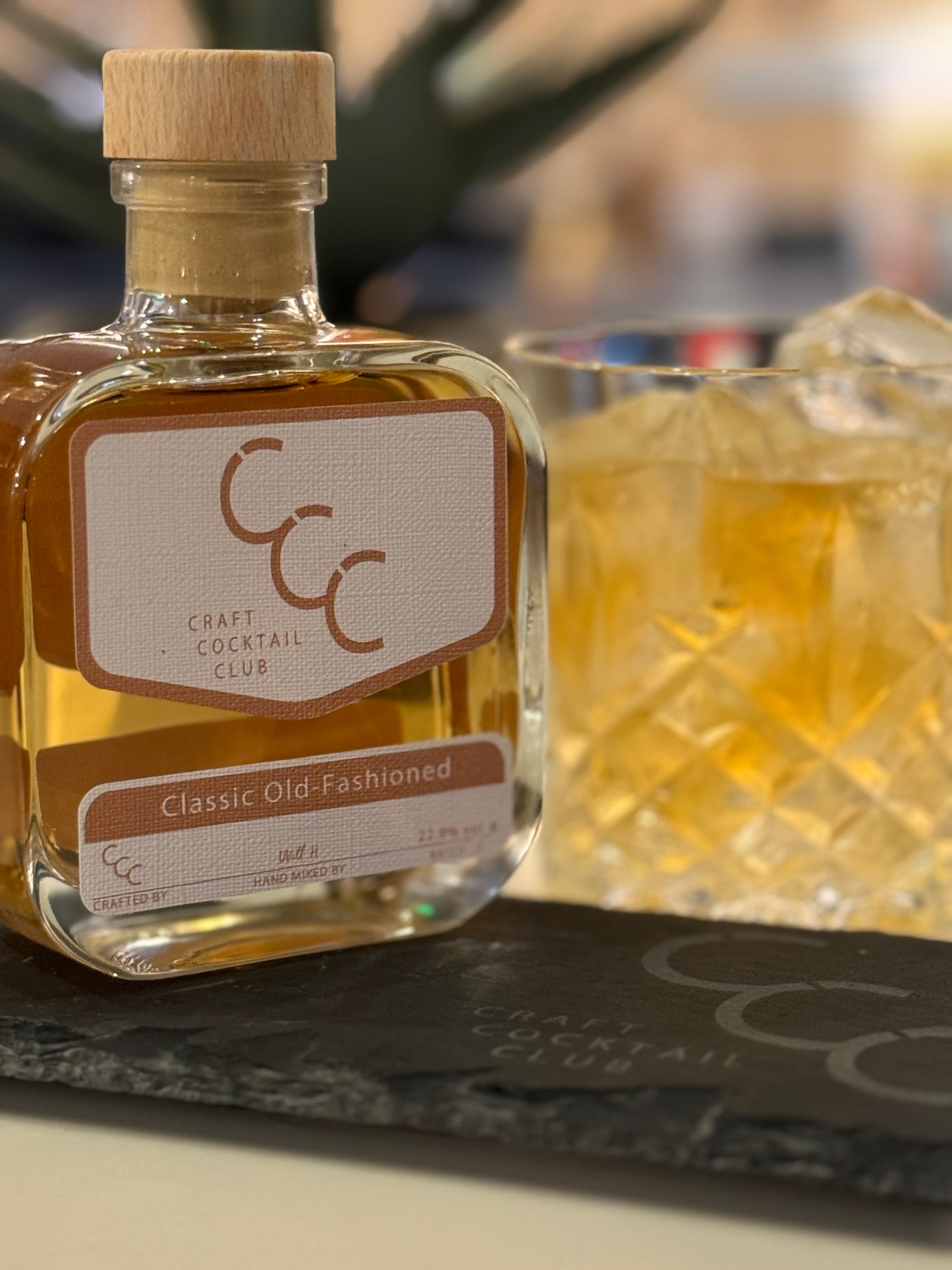 Picture of an old fashioned cocktail in the background, in front of it is a thick square 230ml bottle full of ready to drink cocktail, brown honey coloured on top of a grey slate with and engraving that reads craft cocktail club 