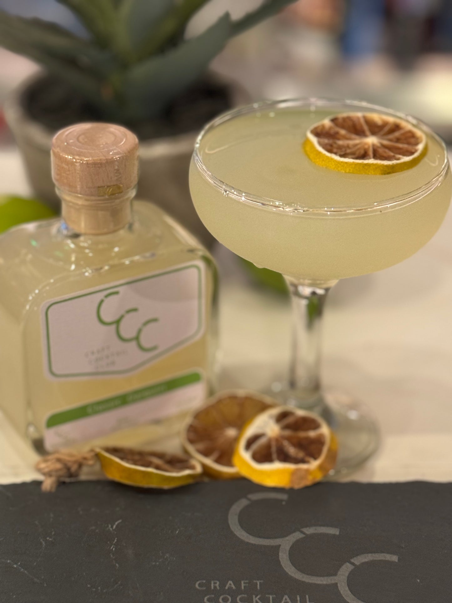 Picture of  a ready made bottle of daiquiri_ on a bar next to a coup glass full of green cocktail, label reads Craft Cocktail Club 