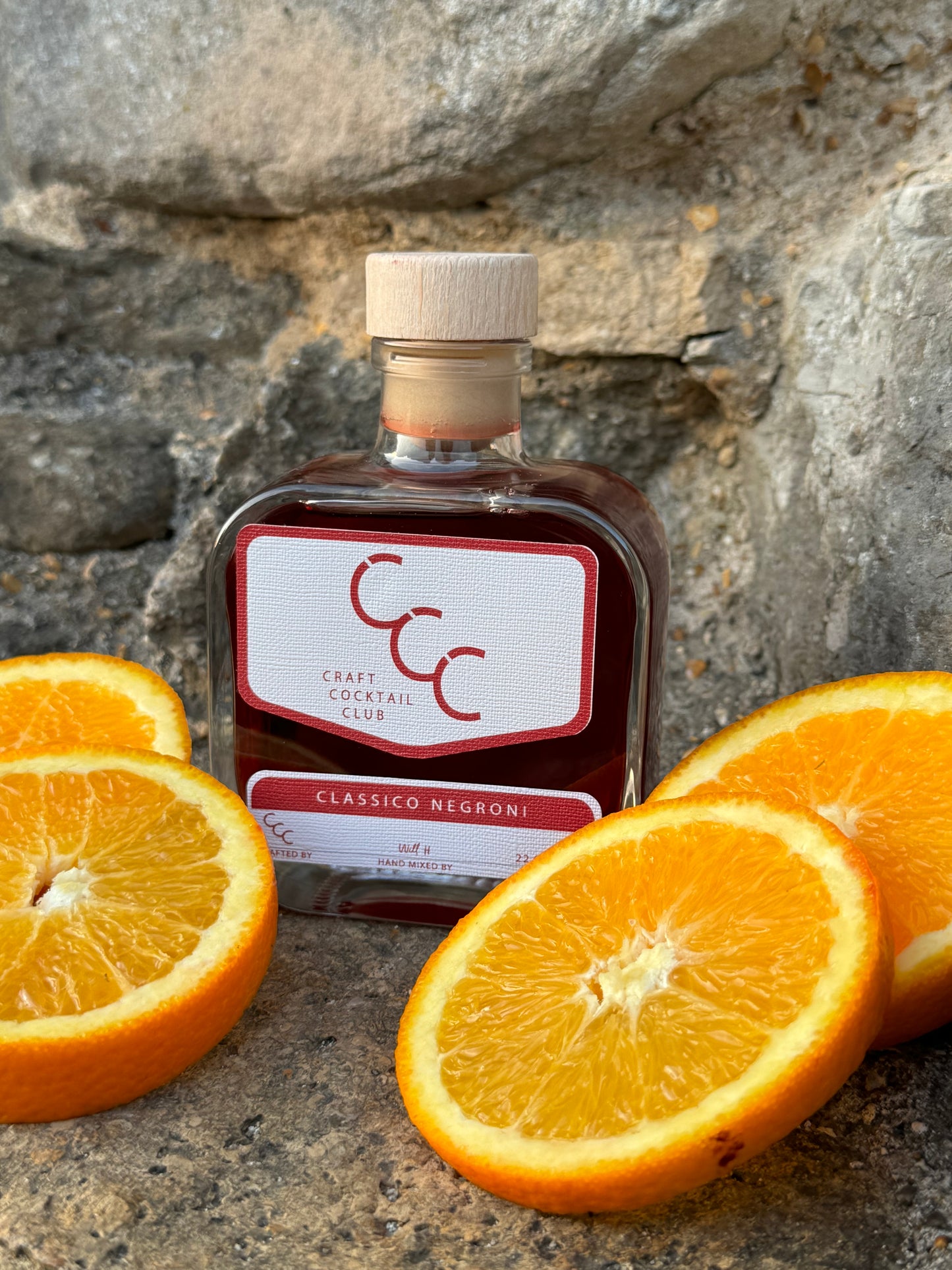 picture of a glass bottle on limestone rock surrounded by cut oranges, in the bottle is a deep red liquid and the label says Craft Cocktail Club - Classic Negroni 